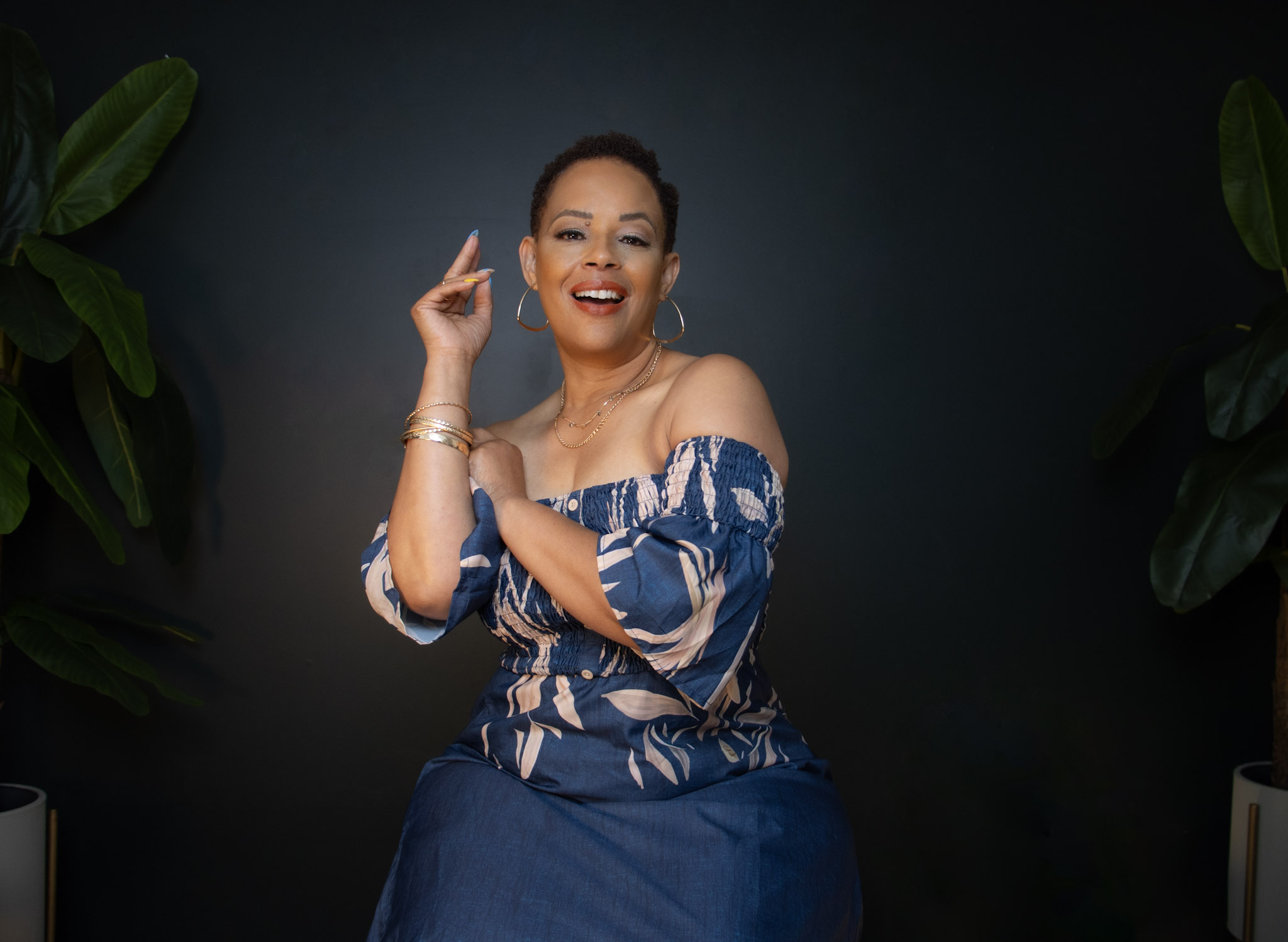 Smiling woman with dark hair in a blue off-shoulder dress, gesturing with her right hand, against a dark background with green plants.
