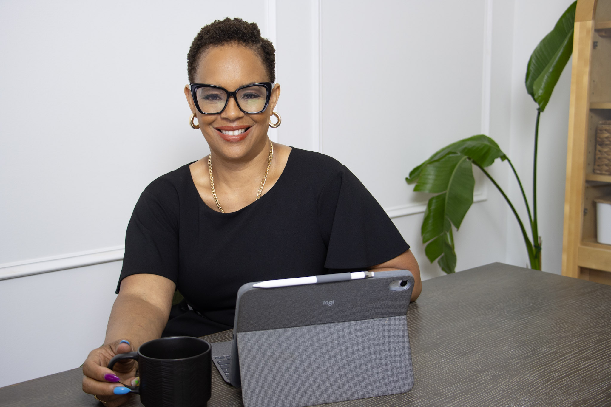 Smiling woman with glasses and earrings sits at a table with a tablet, a black mug, and a plant in the background.