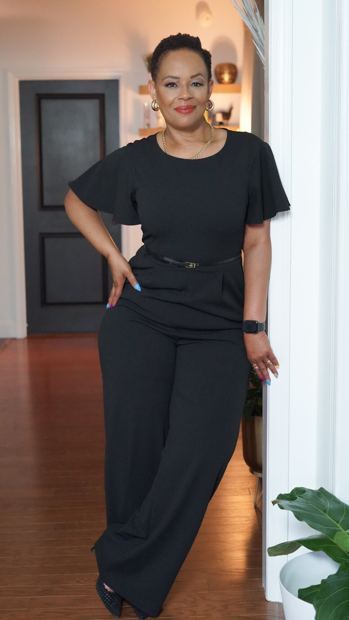 Woman standing indoors, wearing black outfit, smiling, with a door and plant in the background.