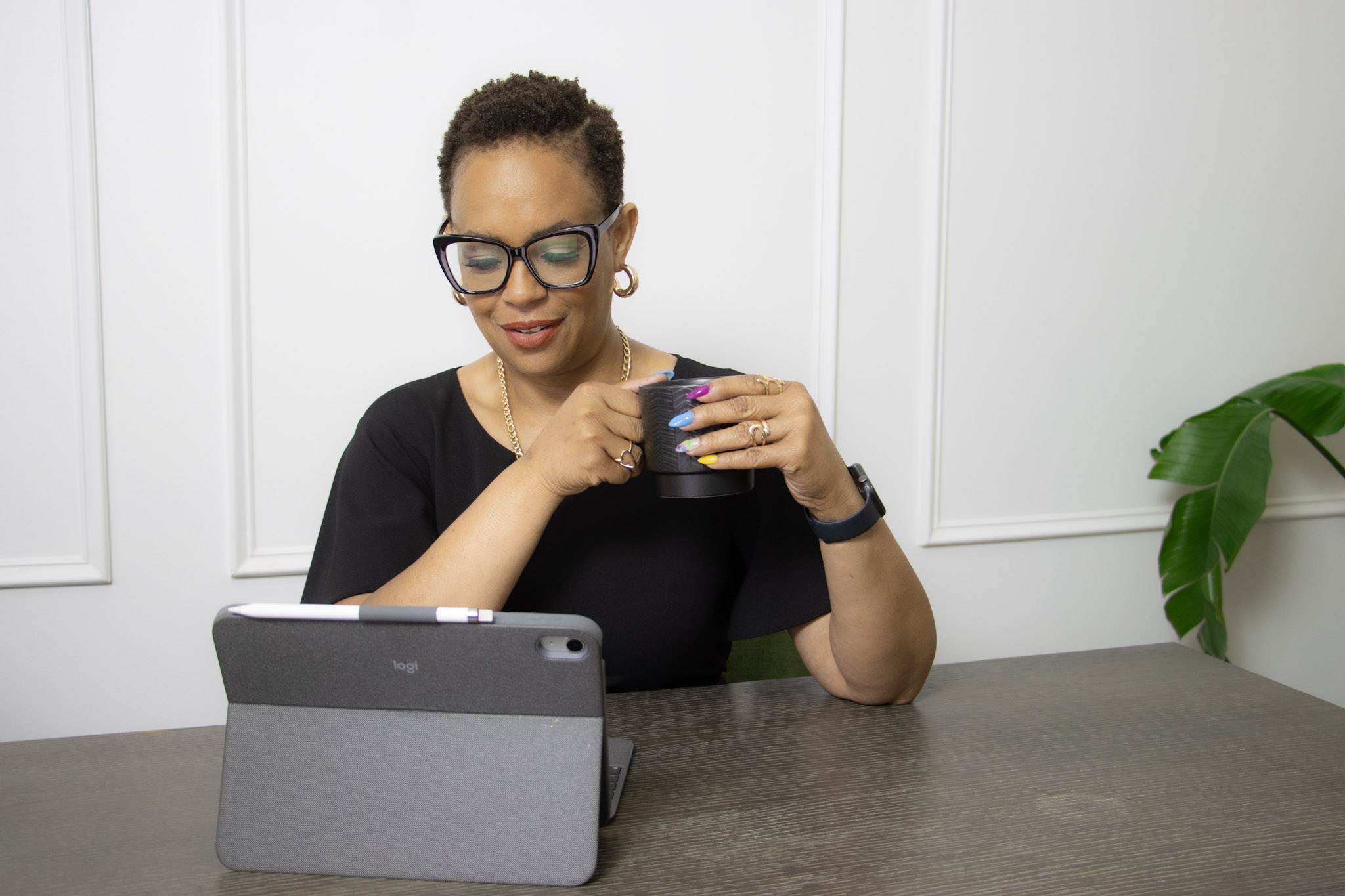 Woman with glasses holding a mug, sitting at a table with a tablet, white wall background, green plant nearby.