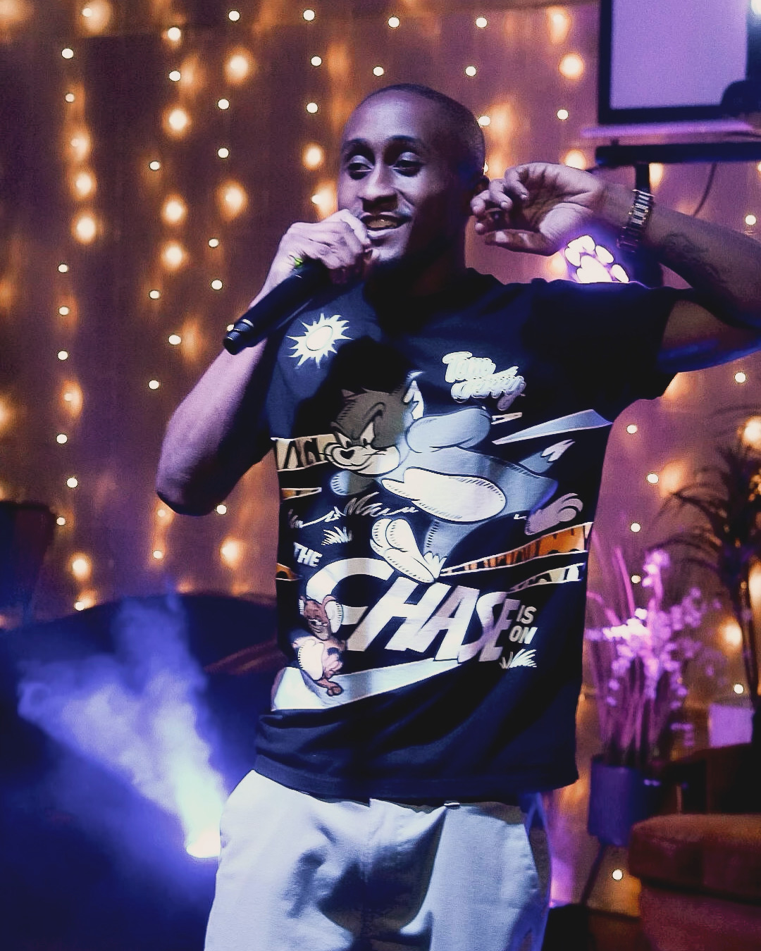 Person holding microphone, smiling, in a decorated indoor setting with string lights and plants, wearing a cartoon graphic T-shirt.