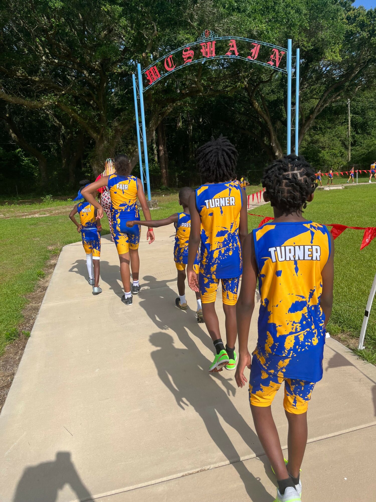 Group of children in yellow and blue sports uniforms standing on a sidewalk, with trees and a banner in the background.