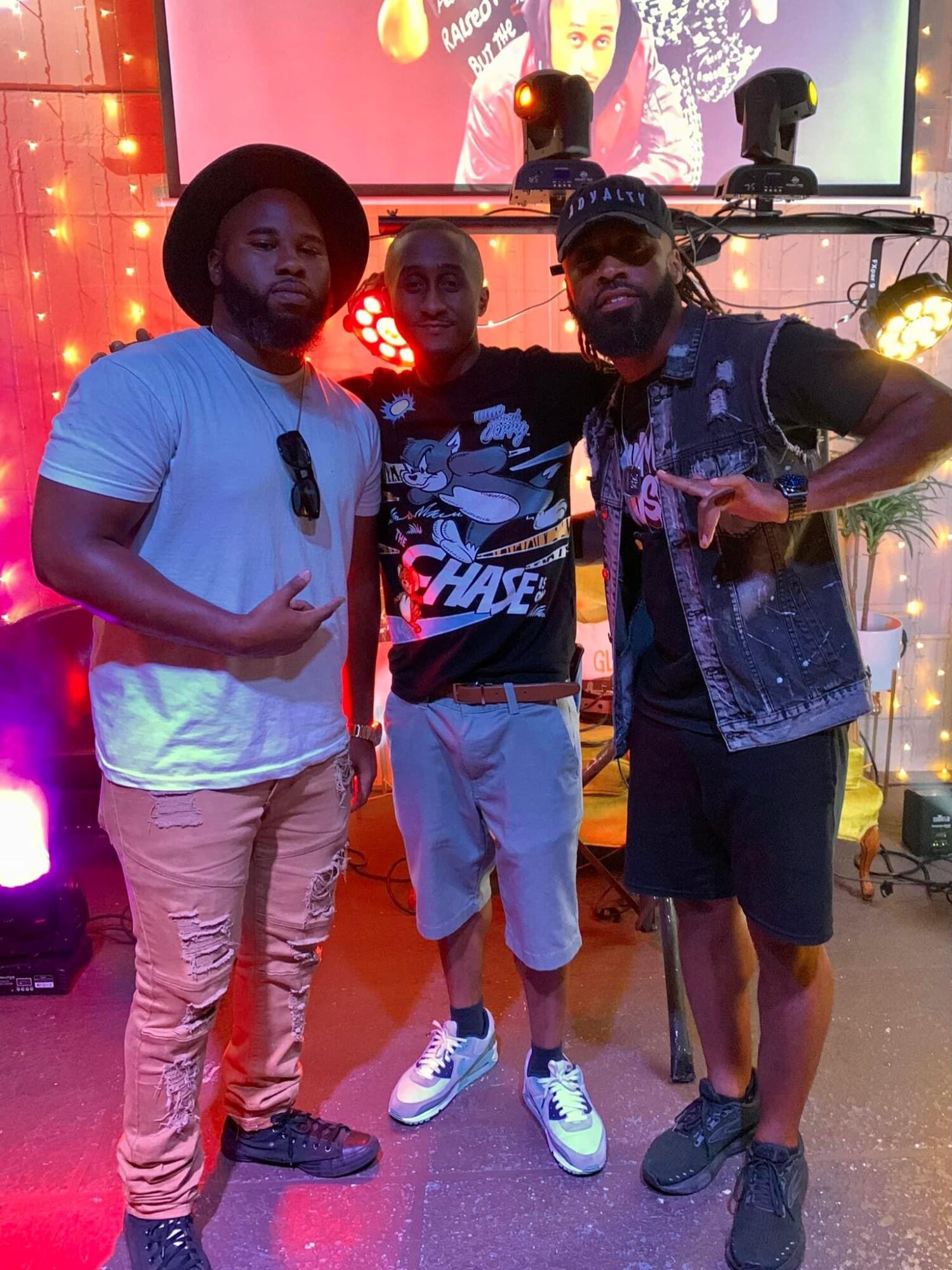 Three men standing together in a colorful indoor setting with stage lights and a screen in the background.