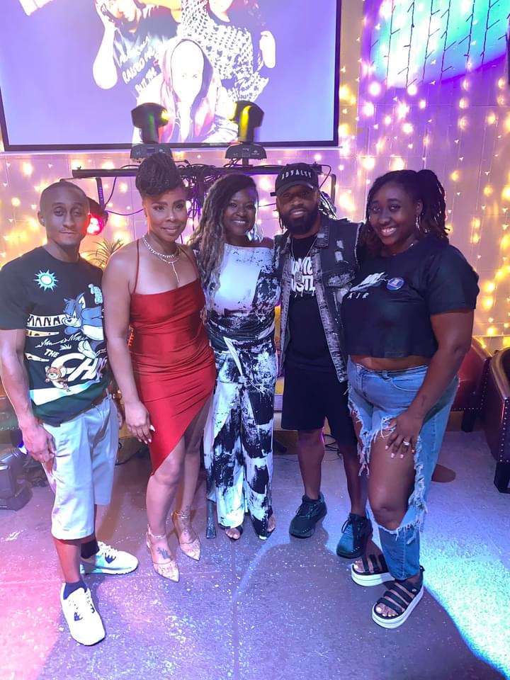 Five people stand together in a colorful, decorated room with a large screen and string lights behind them.