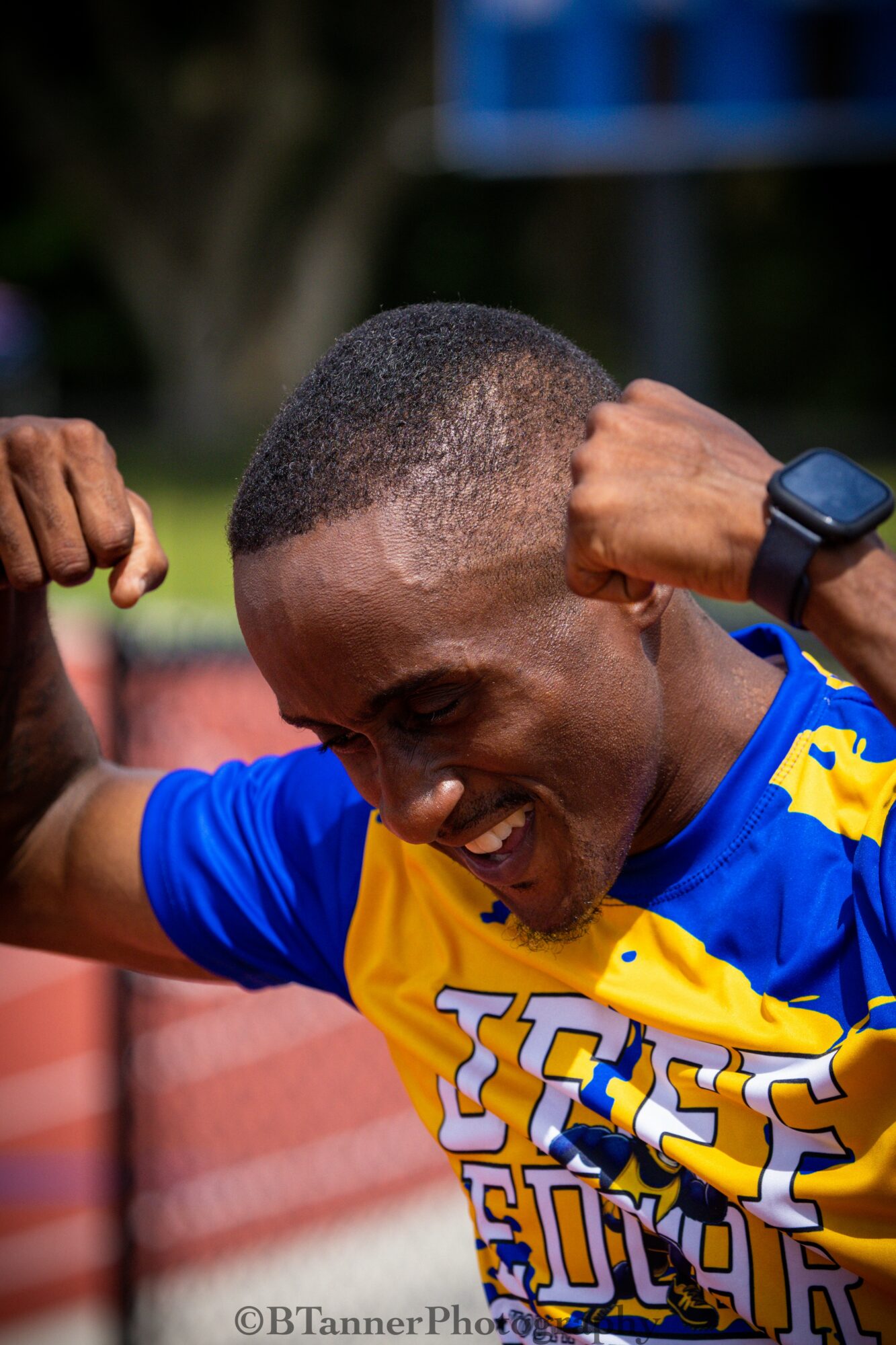 Person smiling and flexing muscles outdoors, wearing a blue and yellow sports jersey and a smartwatch.