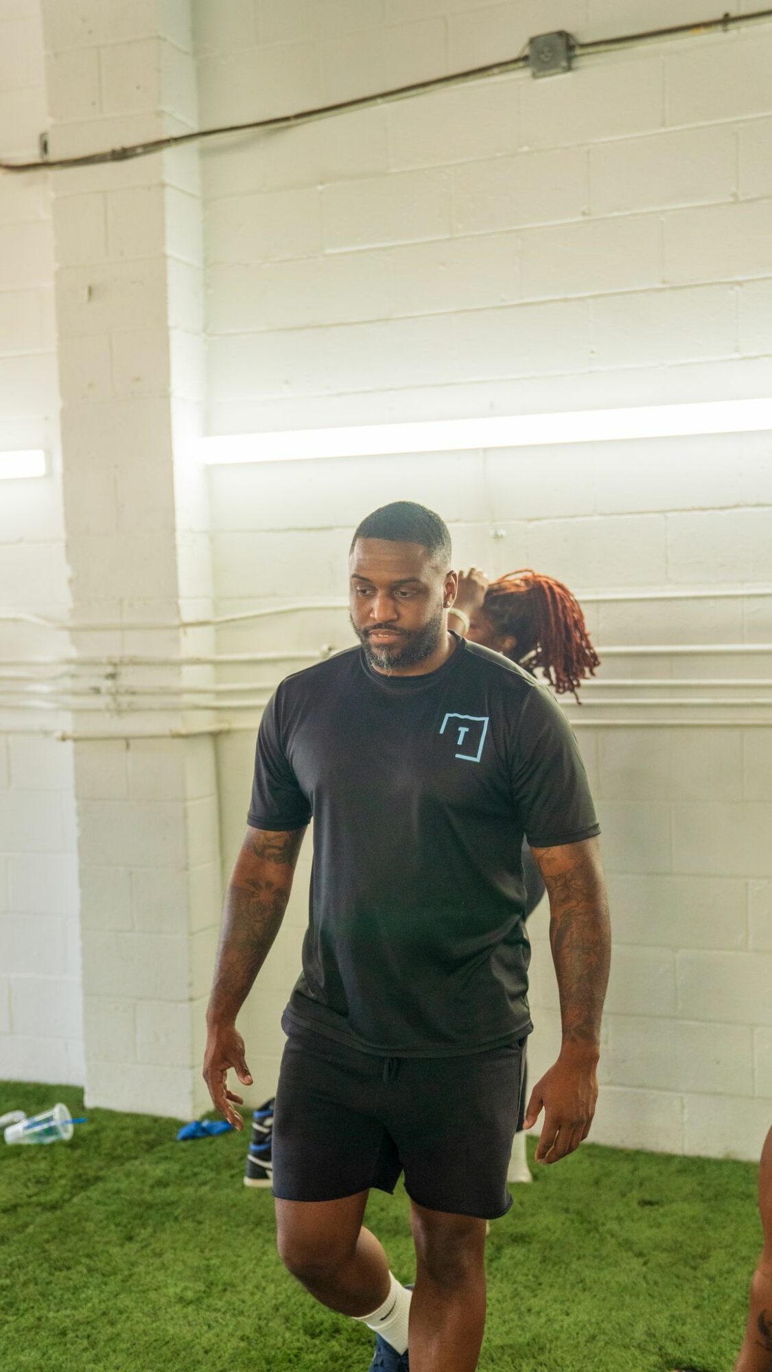 Man with a beard and tattoos wearing a black t-shirt and shorts standing on grass indoors.
