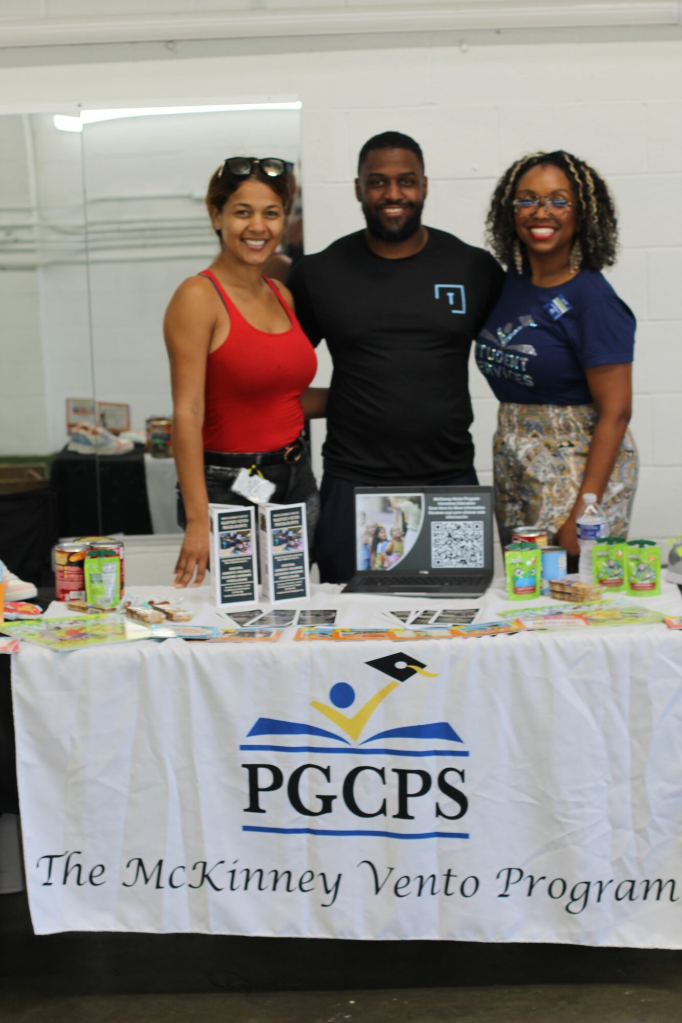 Three people standing behind a table with a PGPS banner, smiling at the camera, in an indoor setting.