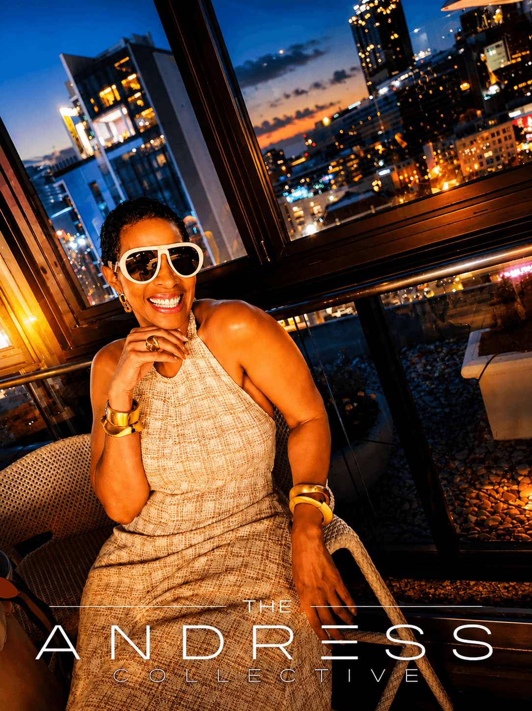 Smiling woman wearing sunglasses and a sleeveless dress sitting by a window with a city skyline at night.