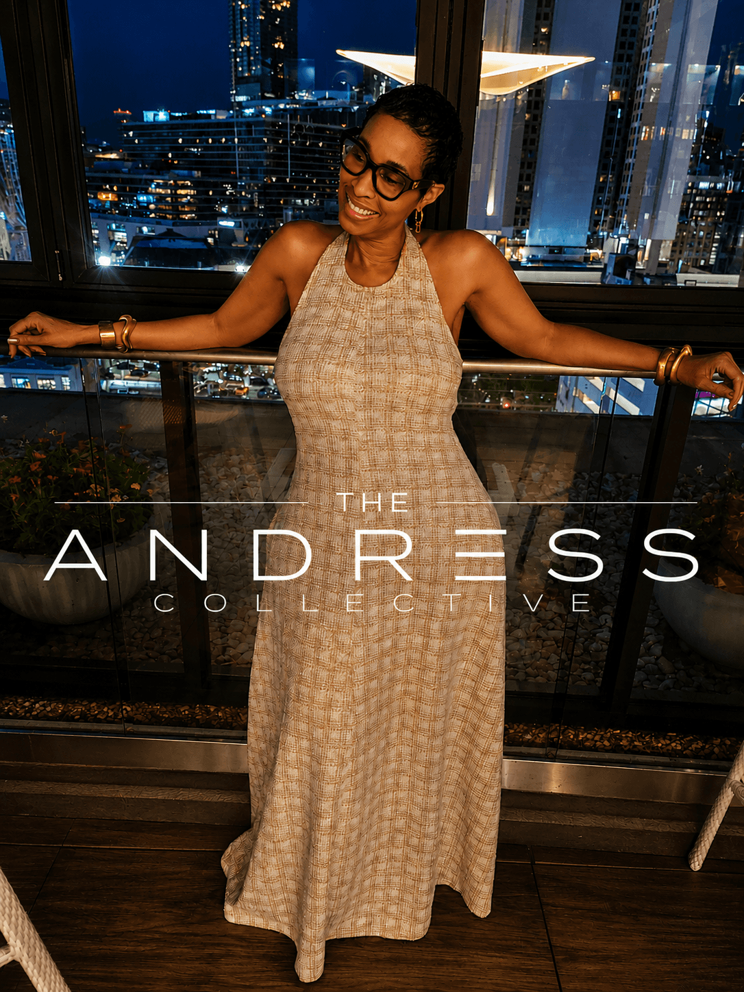Woman in a long dress with arms outstretched, smiling, city skyline at night behind her.