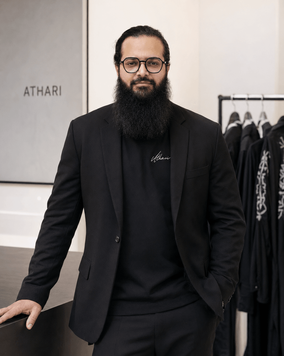 Man with glasses and a beard wearing a black blazer and shirt, standing indoors near clothing racks.