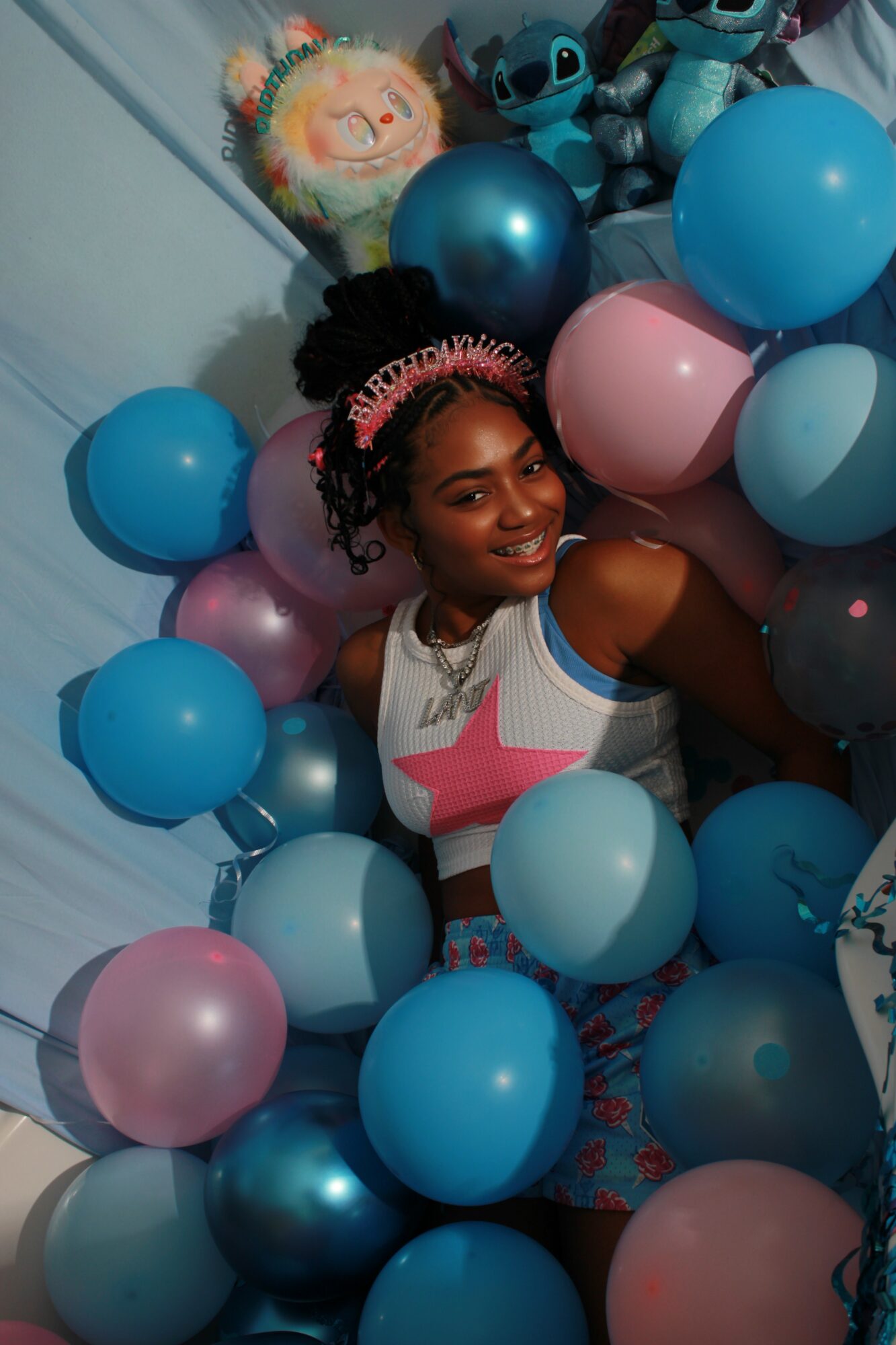 Smiling woman surrounded by pink and blue balloons, with Stitch plush toys and a colorful plush in the background.