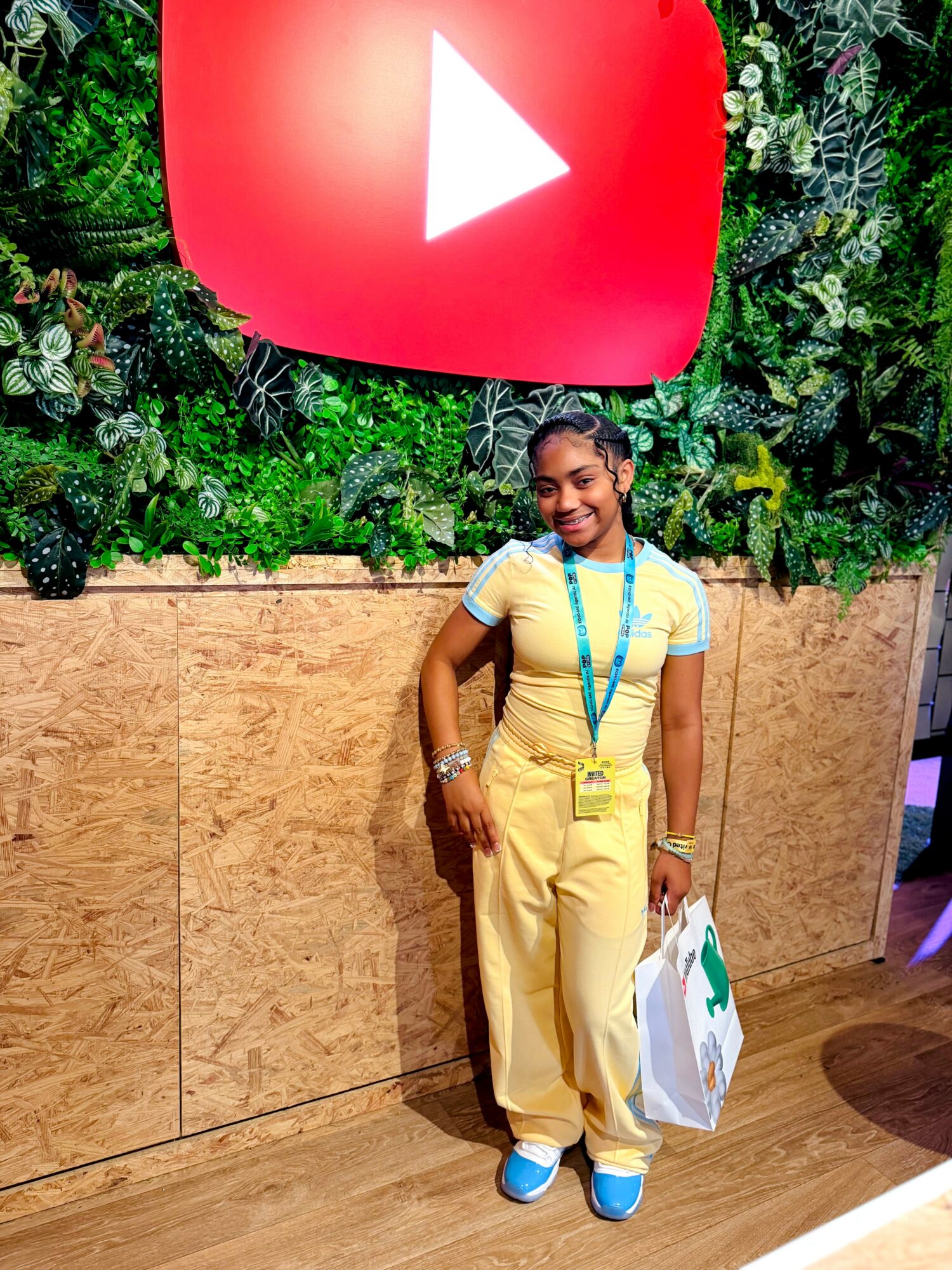 Young girl standing in front of a large YouTube logo with greenery background, holding a shopping bag, smiling, wearing yellow outfit and blue shoes.