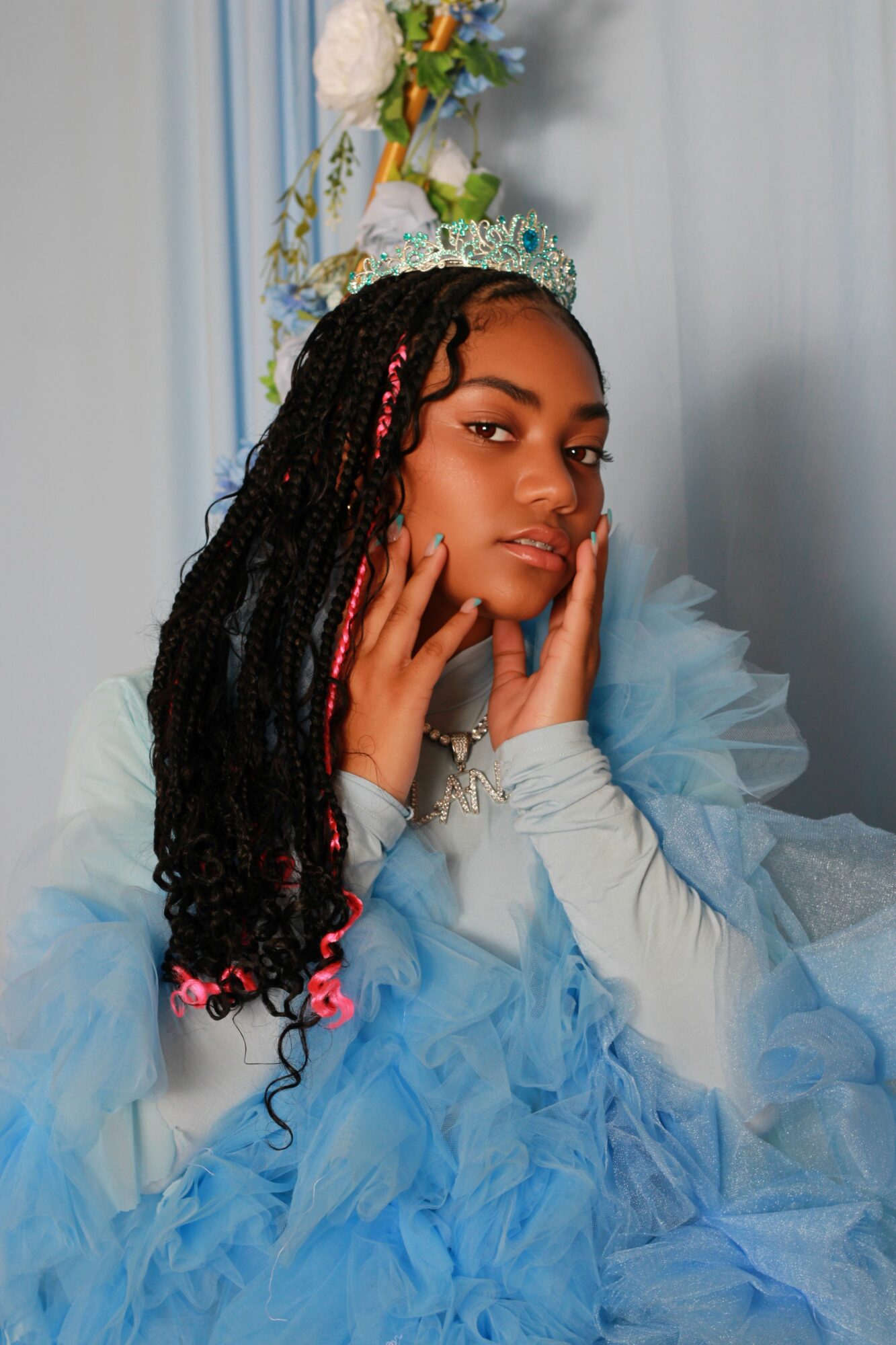 Young woman with long braided hair wearing a blue dress and crown, posing with hands near face, against light background.