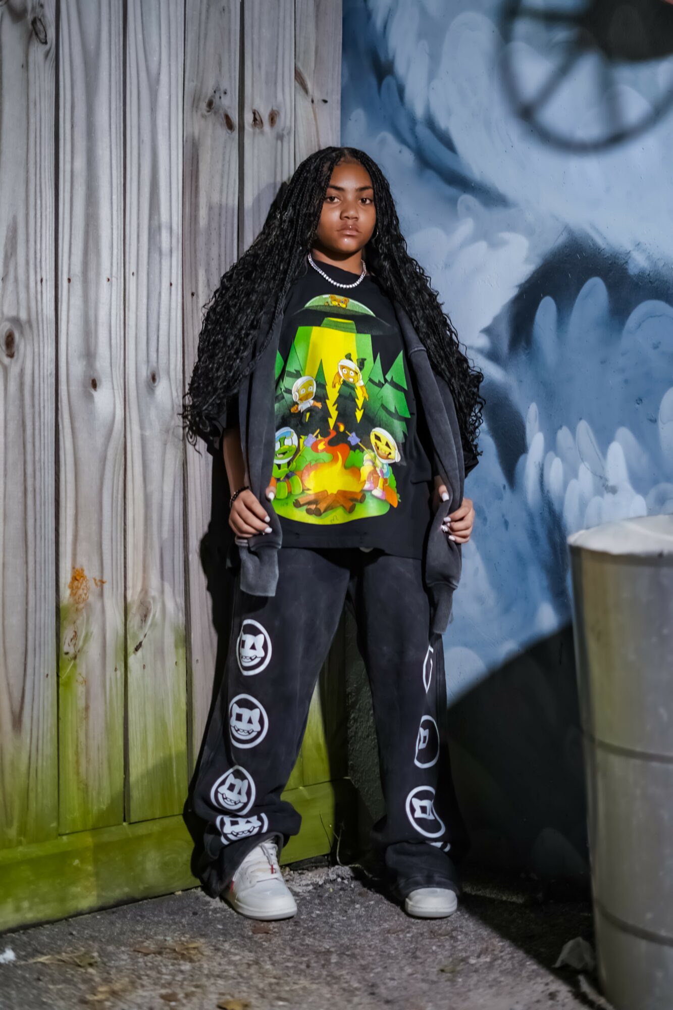 Young woman with long curly hair stands against a wooden wall and a blue mural, wearing a colorful graphic t-shirt and black pants with smiley face patches.