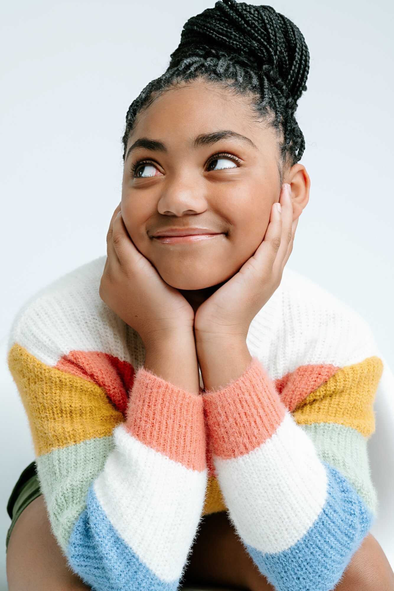 Young girl with braided hair resting her face in her hands, smiling, wearing a colorful striped sweater.