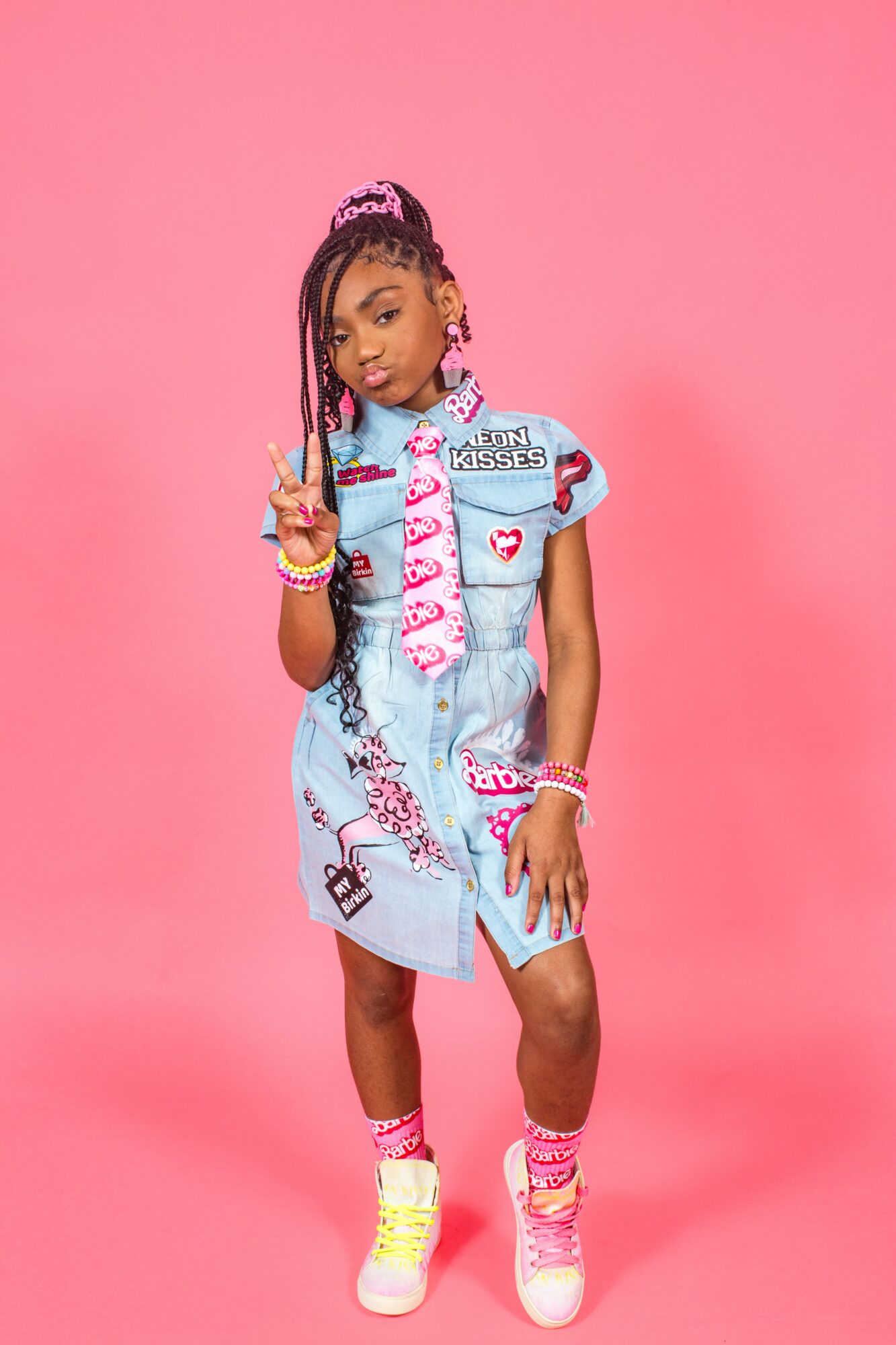 Young girl with braided hair and colorful accessories poses against pink background, wearing a denim dress with patches and pink sneakers.
