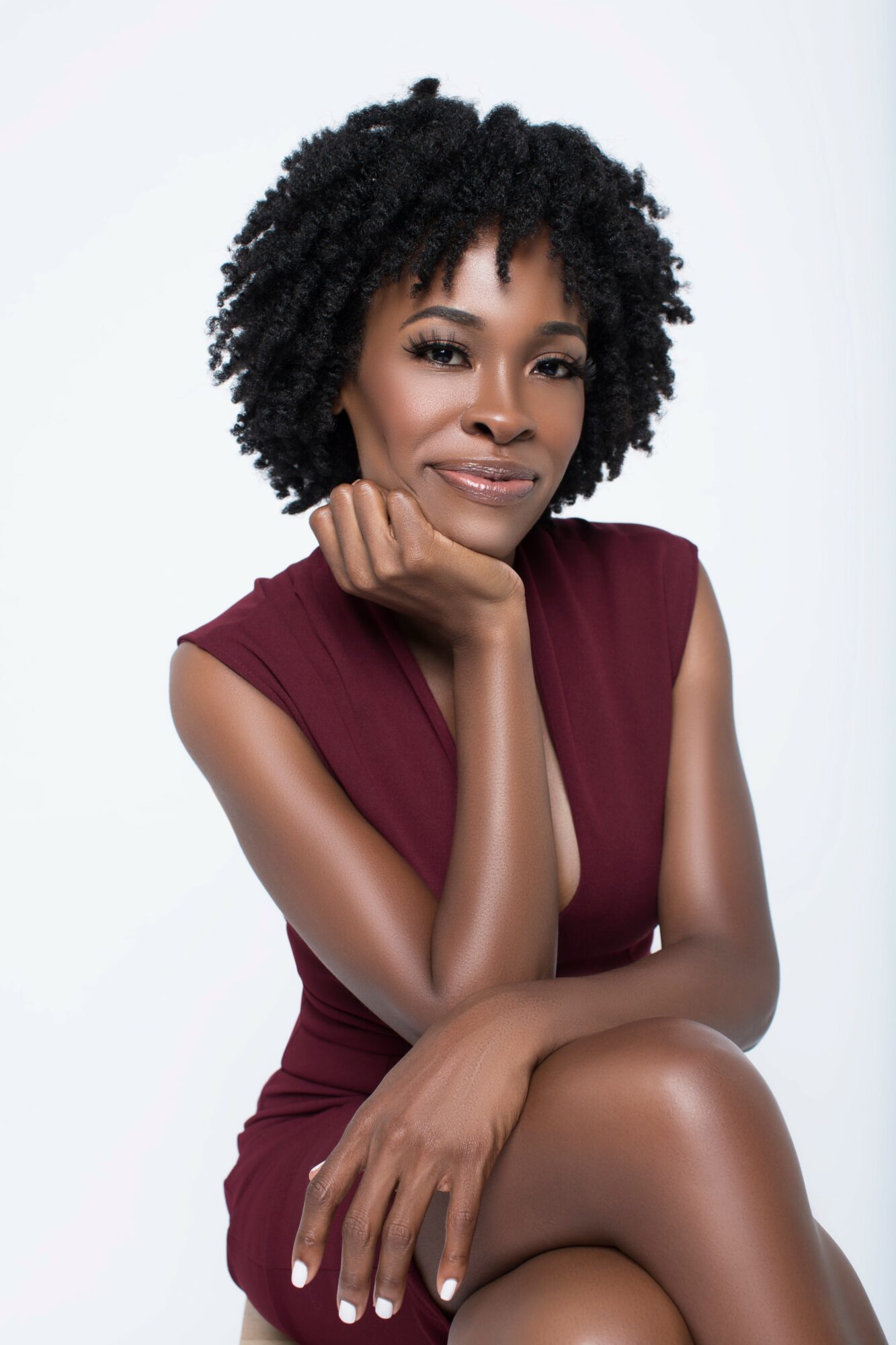 Portrait of a woman with natural curly hair, wearing a sleeveless maroon dress, resting her chin on her hand, smiling.