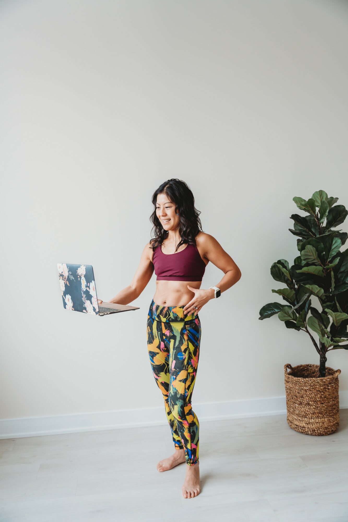 Woman standing barefoot in a room with a laptop, wearing colorful pants and a maroon top, next to a potted plant.