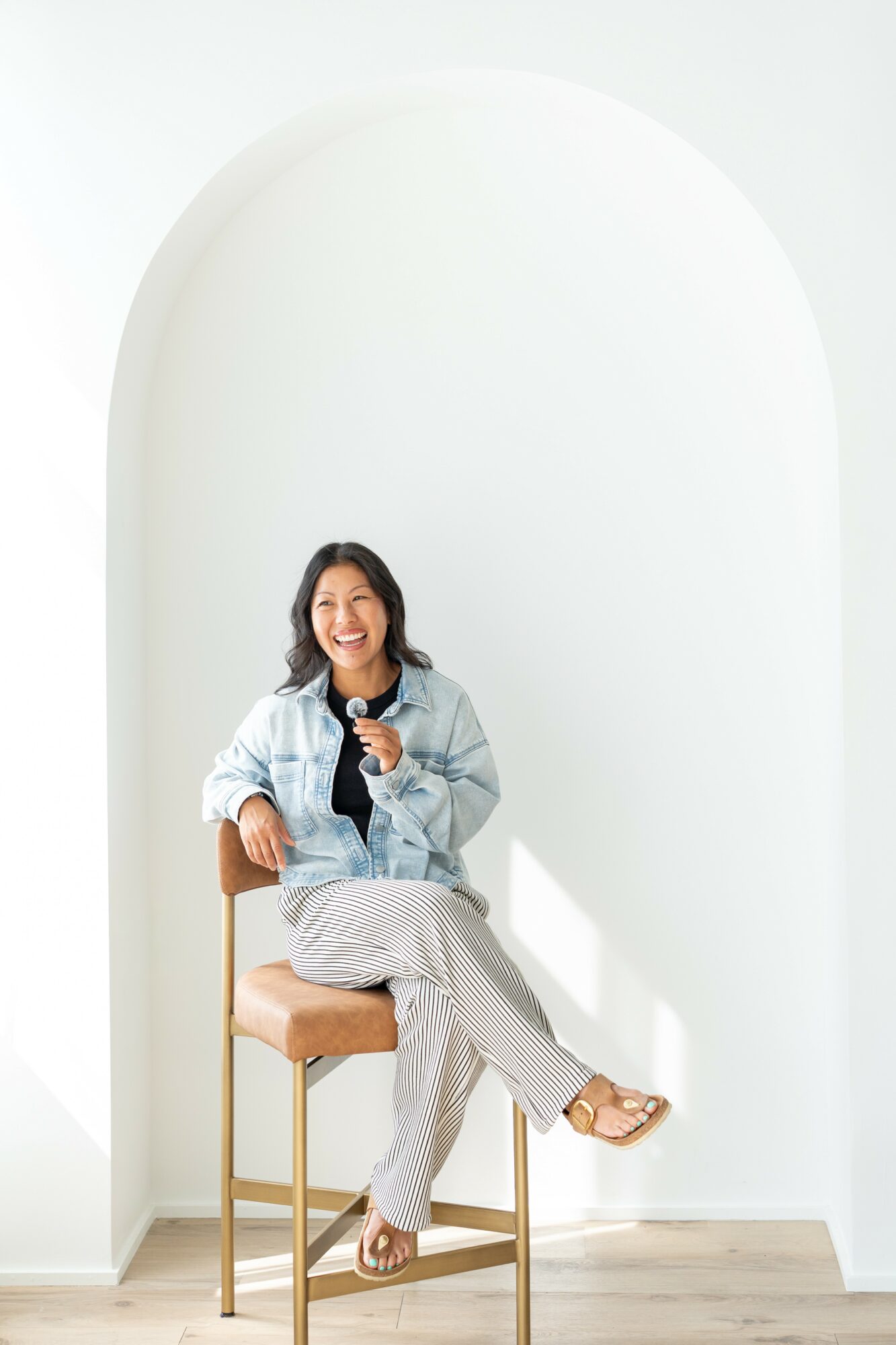 Woman sitting on a chair, smiling, holding a microphone, in a bright room with an arched wall.