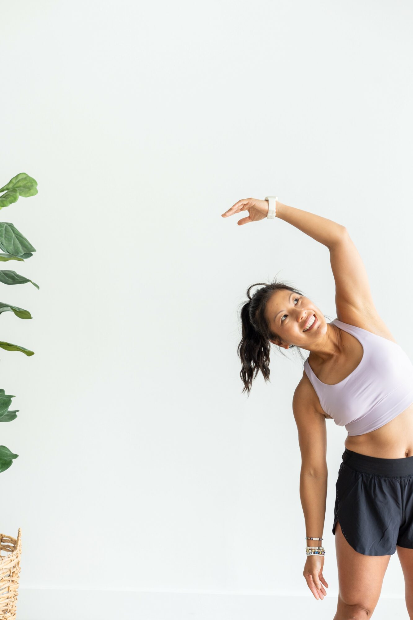 Woman stretching with her arm raised, wearing a white sports bra and black shorts, standing next to a plant, smiling.