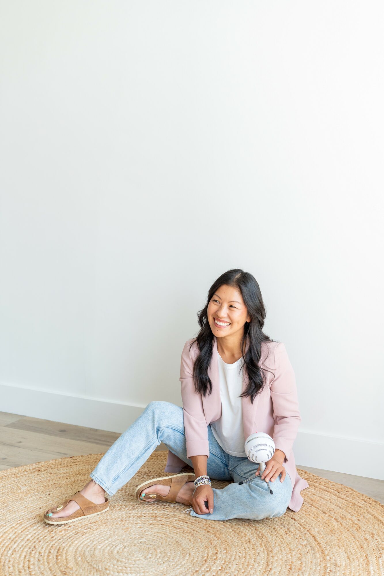 Young woman sitting on a round rug, smiling, against a plain white wall, wearing casual clothes.