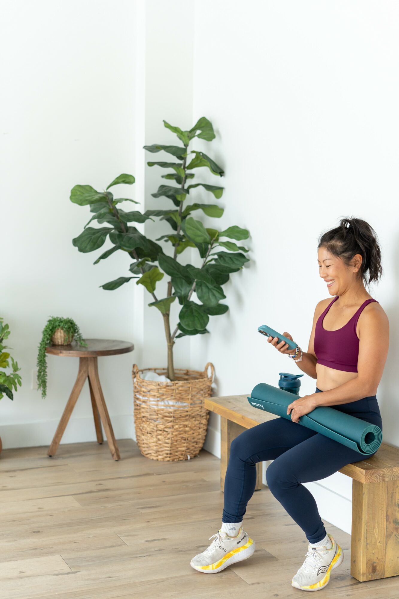 Woman sitting on a wooden bench, holding a phone, with a large potted plant and small table in a bright room.