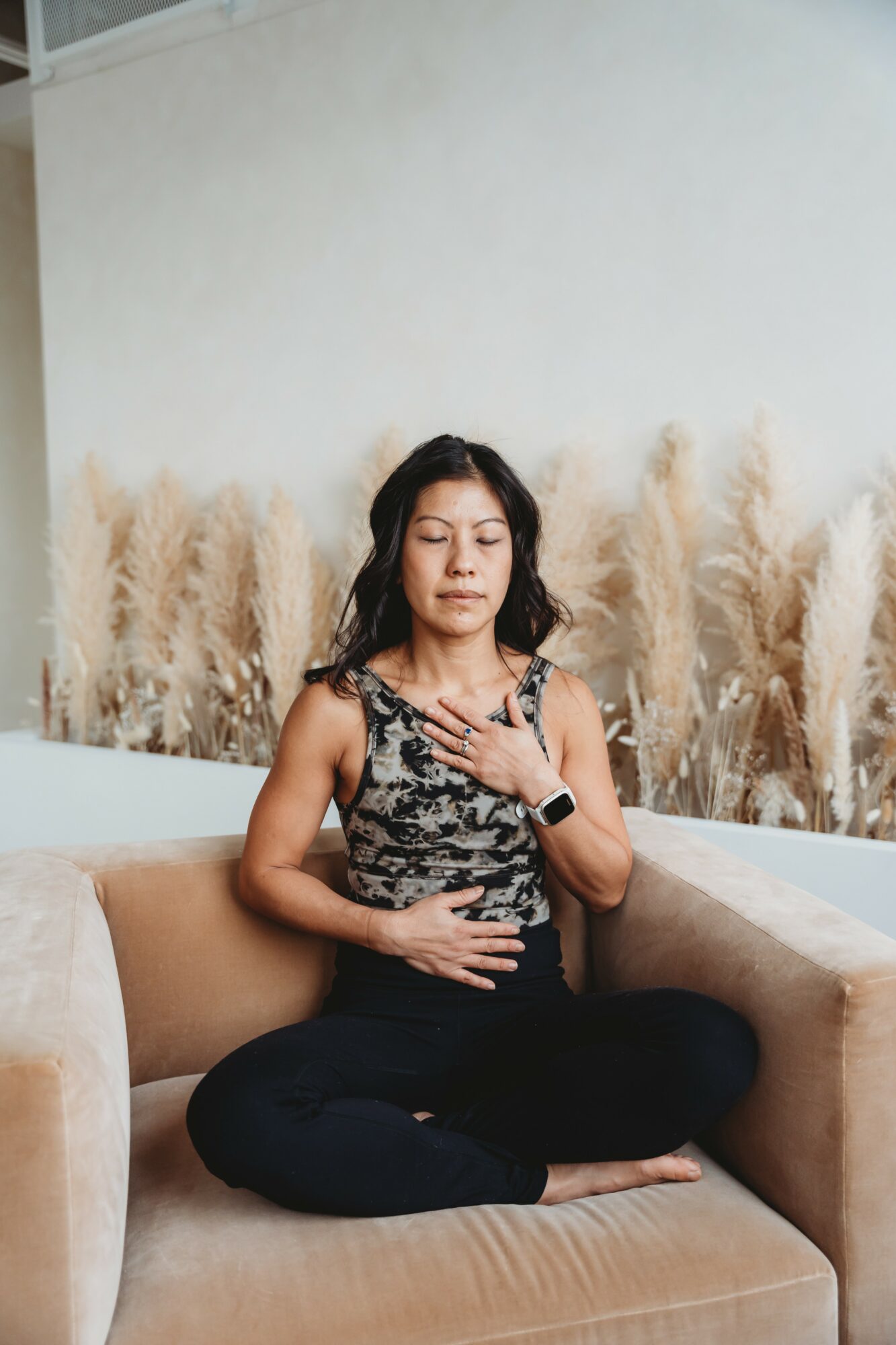 Woman sitting cross-legged on a sofa with eyes closed, hand on chest, in a room with tall plants behind her.