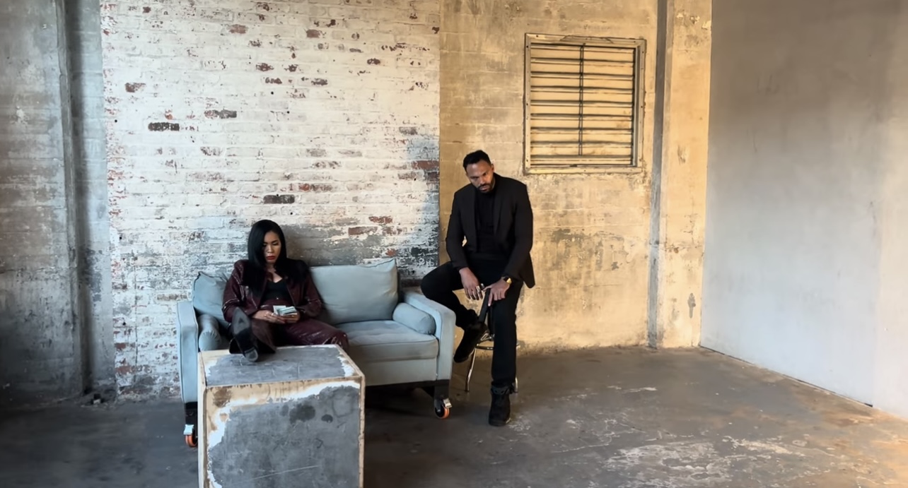 Two people in a room with a brick wall, one sitting on a sofa and the other standing nearby.
