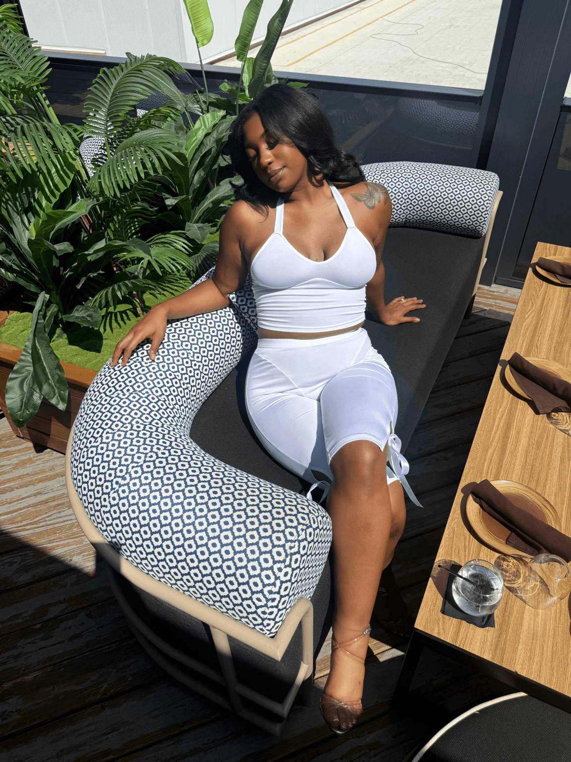 Woman in white outfit sitting on a patterned chair near a window with plants, sunlight streaming in.