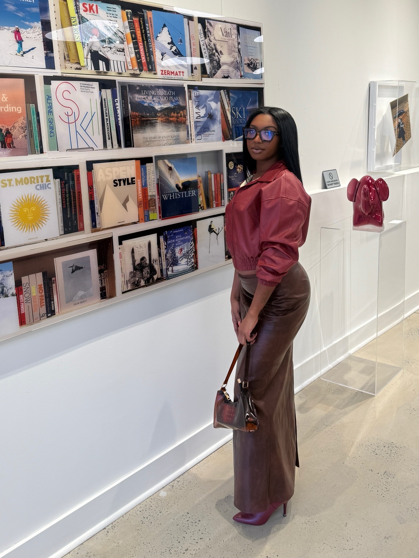Woman with glasses and long dark hair stands in art gallery, looking at books on display shelf, holding a handbag.