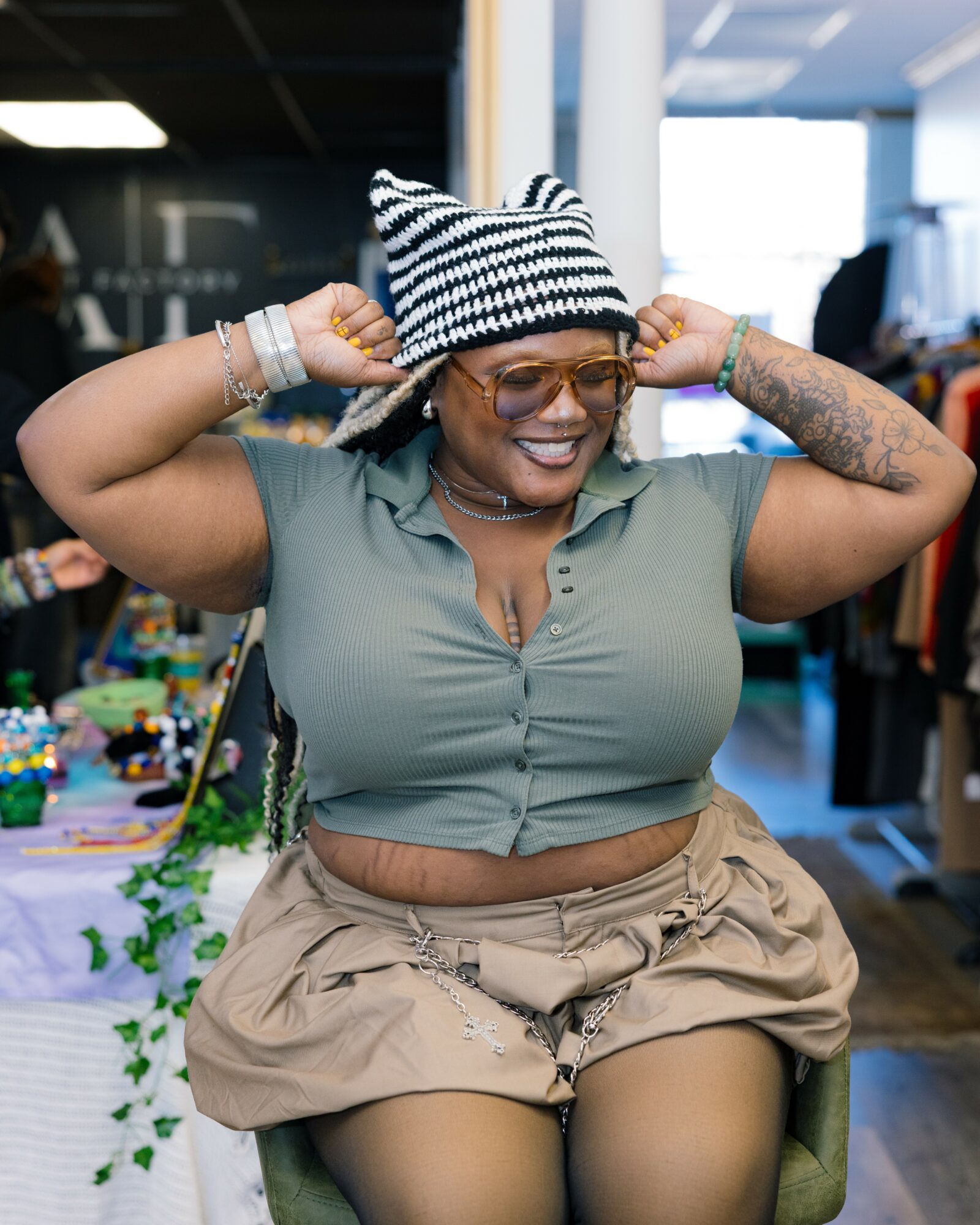 Woman smiling, wearing striped hat, glasses, and casual clothing, adjusting her hat with both hands in a store or market setting.