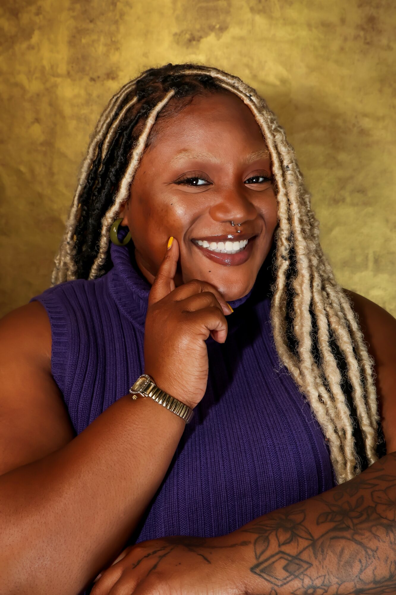 Smiling woman with long blonde dreadlocks wearing a purple sleeveless top, resting her chin on her hand, against a yellow background.