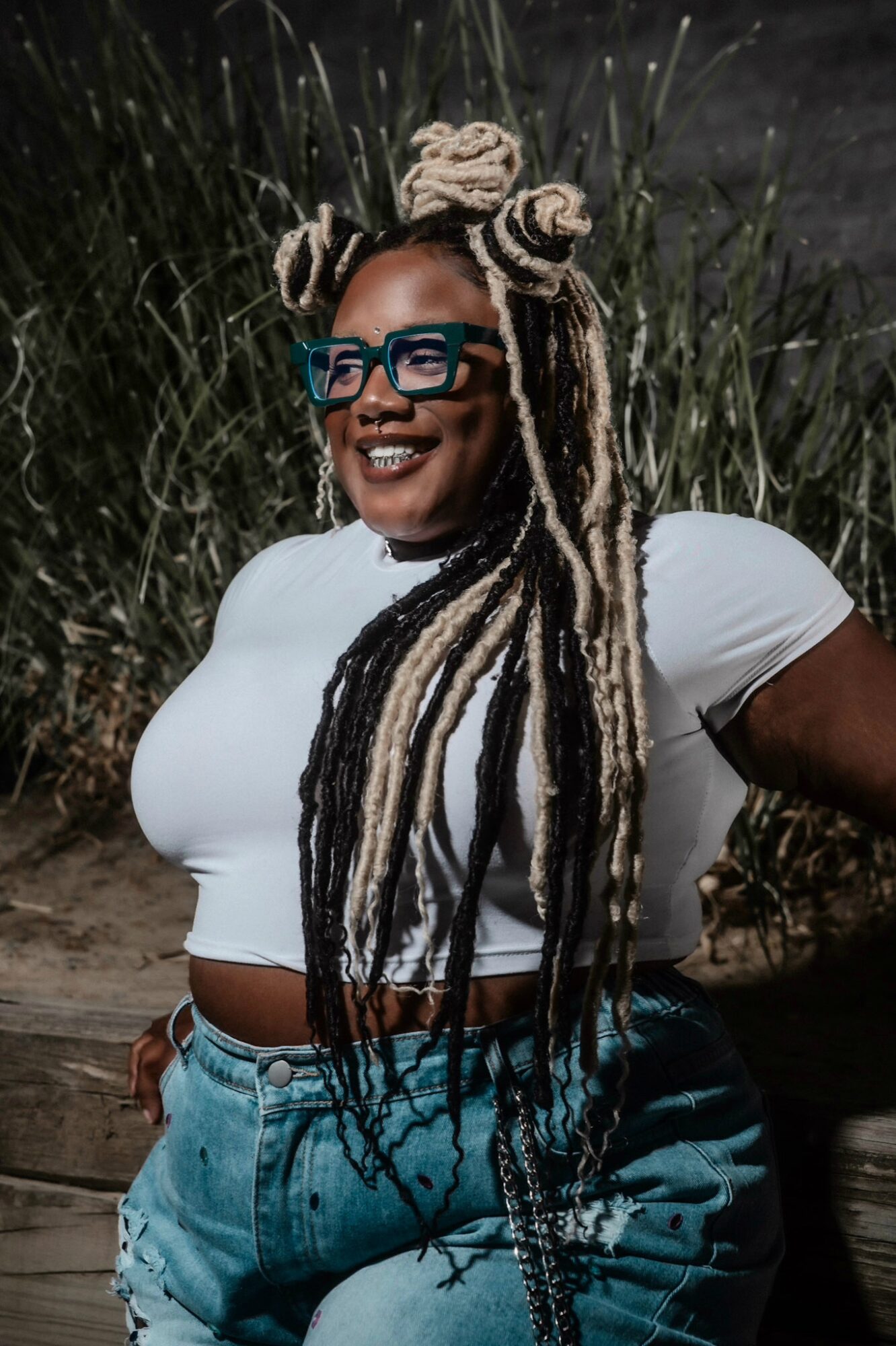 Smiling woman with long, styled dreadlocks, wearing glasses and a white crop top outdoors.