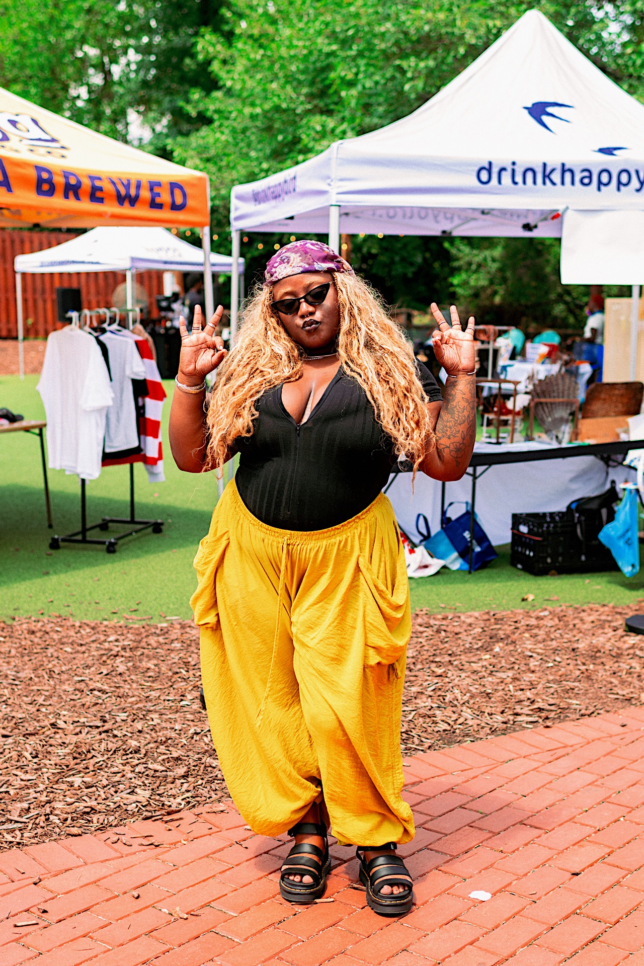 Woman with long curly hair, sunglasses, black top, yellow pants, standing outdoors with tents and trees in background.