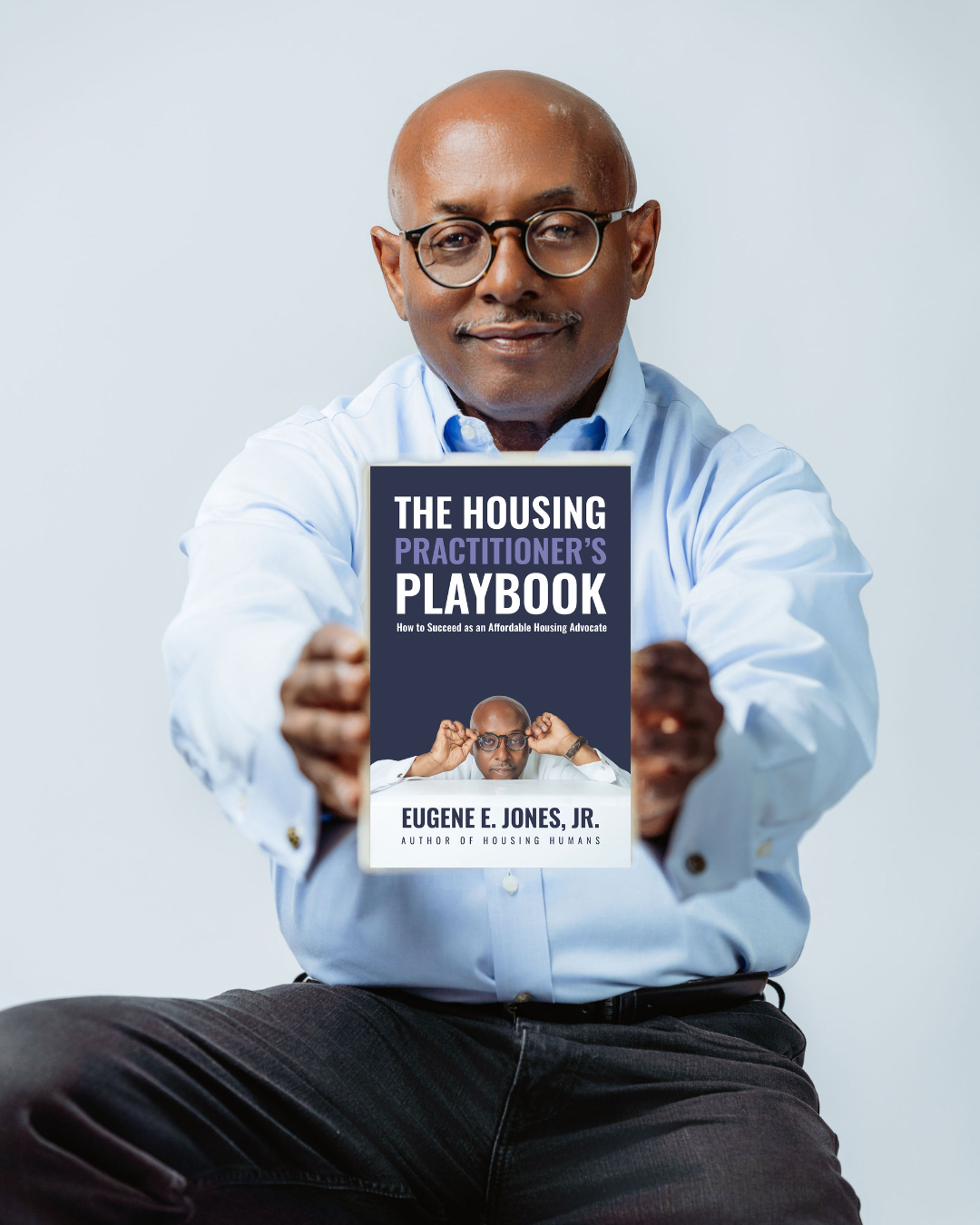 Man holding a book titled 'The Housing Practitioner’s Playbook' smiling, wearing glasses and a light blue shirt, seated against a light background.