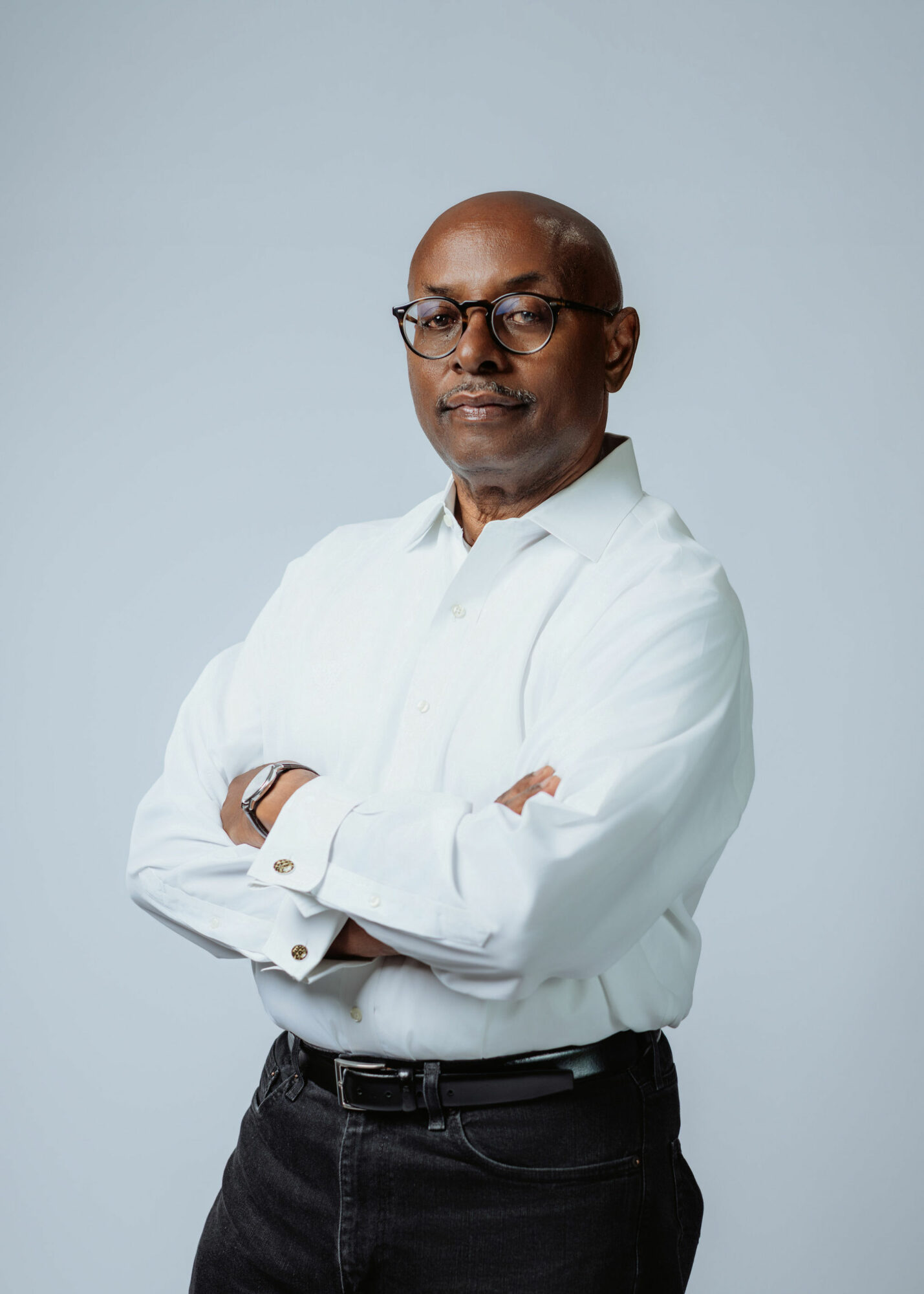 Man with glasses and a white shirt standing with arms crossed against a light background.