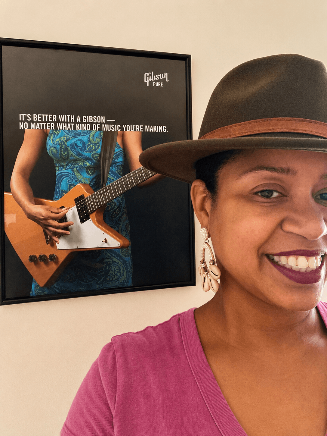 Smiling woman wearing a hat and earrings, with a framed poster of a person playing guitar in the background.