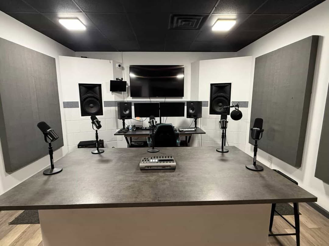 Recording studio with speakers, microphones, and a mixing console on a large table, with a TV and soundproofing panels in the background.