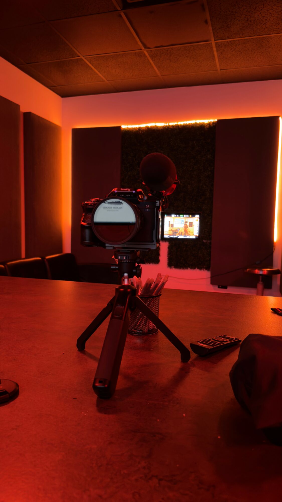 Camera on tripod facing a room with a dark wall and vertical light strips, with a person visible on a screen.