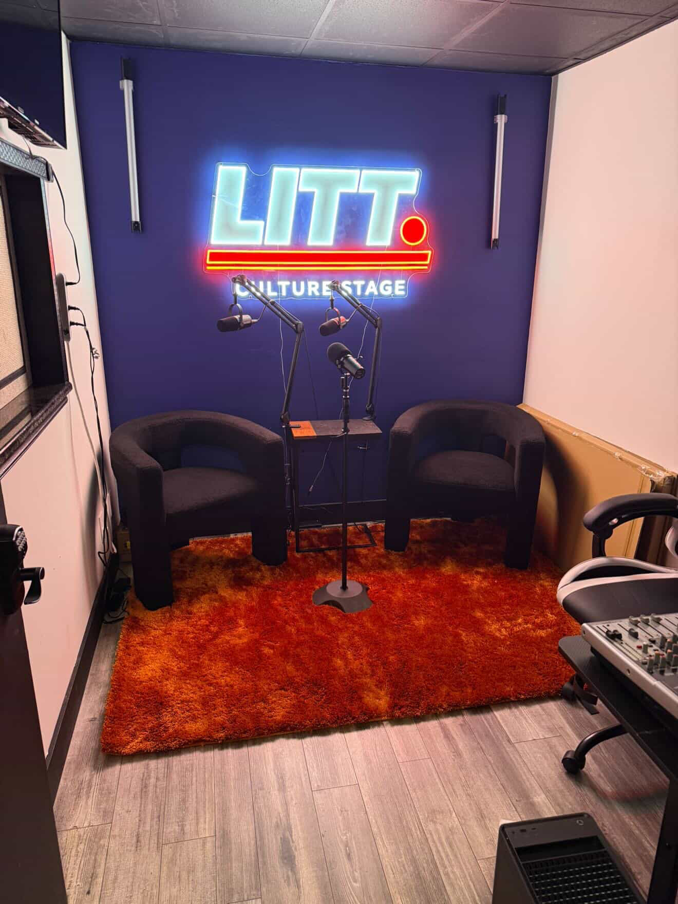 Stage setup with two black chairs, a microphone stand, a neon sign reading 'LITT', and a red rug.
