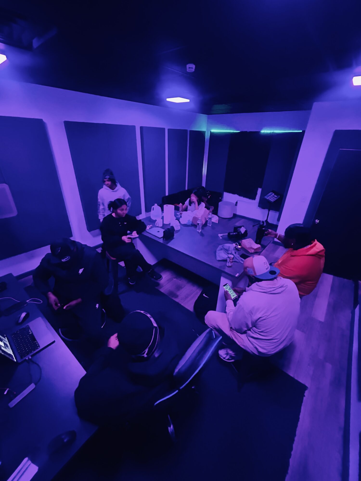 Group of people in a dimly lit room with blue and purple lighting, sitting around a table with electronic equipment.