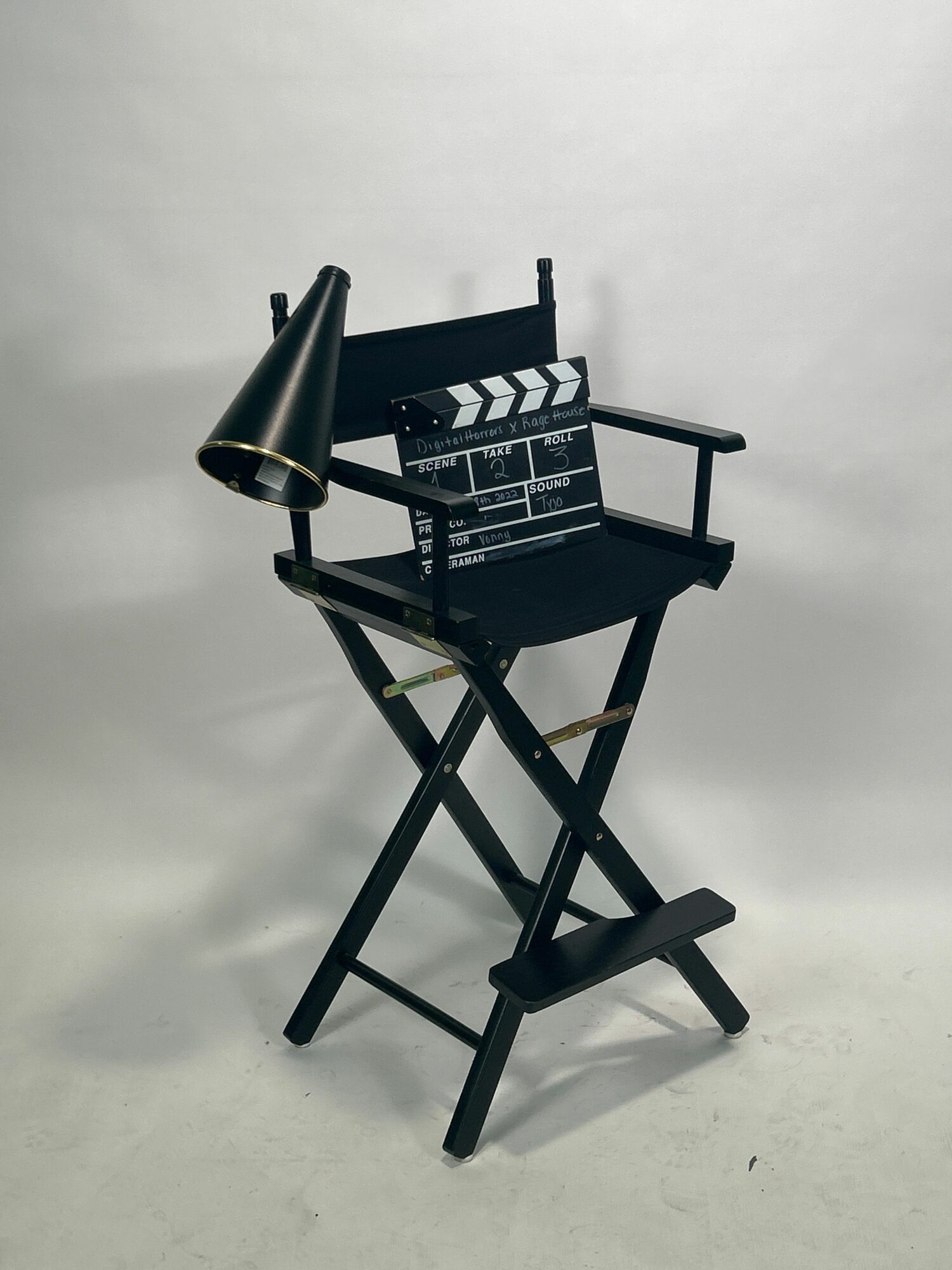 Black chair with a small black table attached, a black cone-shaped object, and a black stool on a white wall background.