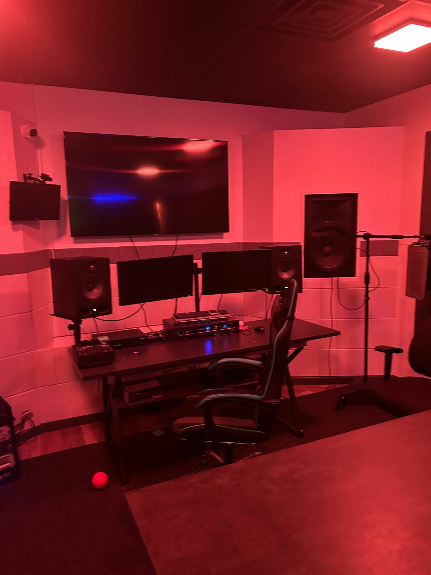 Music studio with a large monitor, speakers, and DJ equipment in a room with pink lighting.