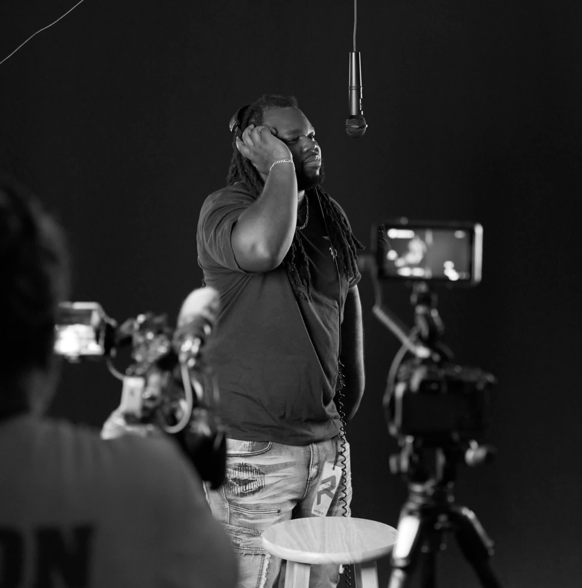 Man with long hair and beard in a recording studio, adjusting headphones, with camera and microphone, in black and white.