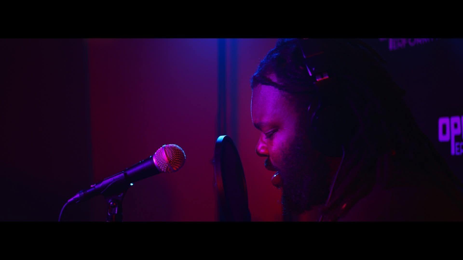 Person singing into a microphone in a dimly lit setting with purple and blue lighting.