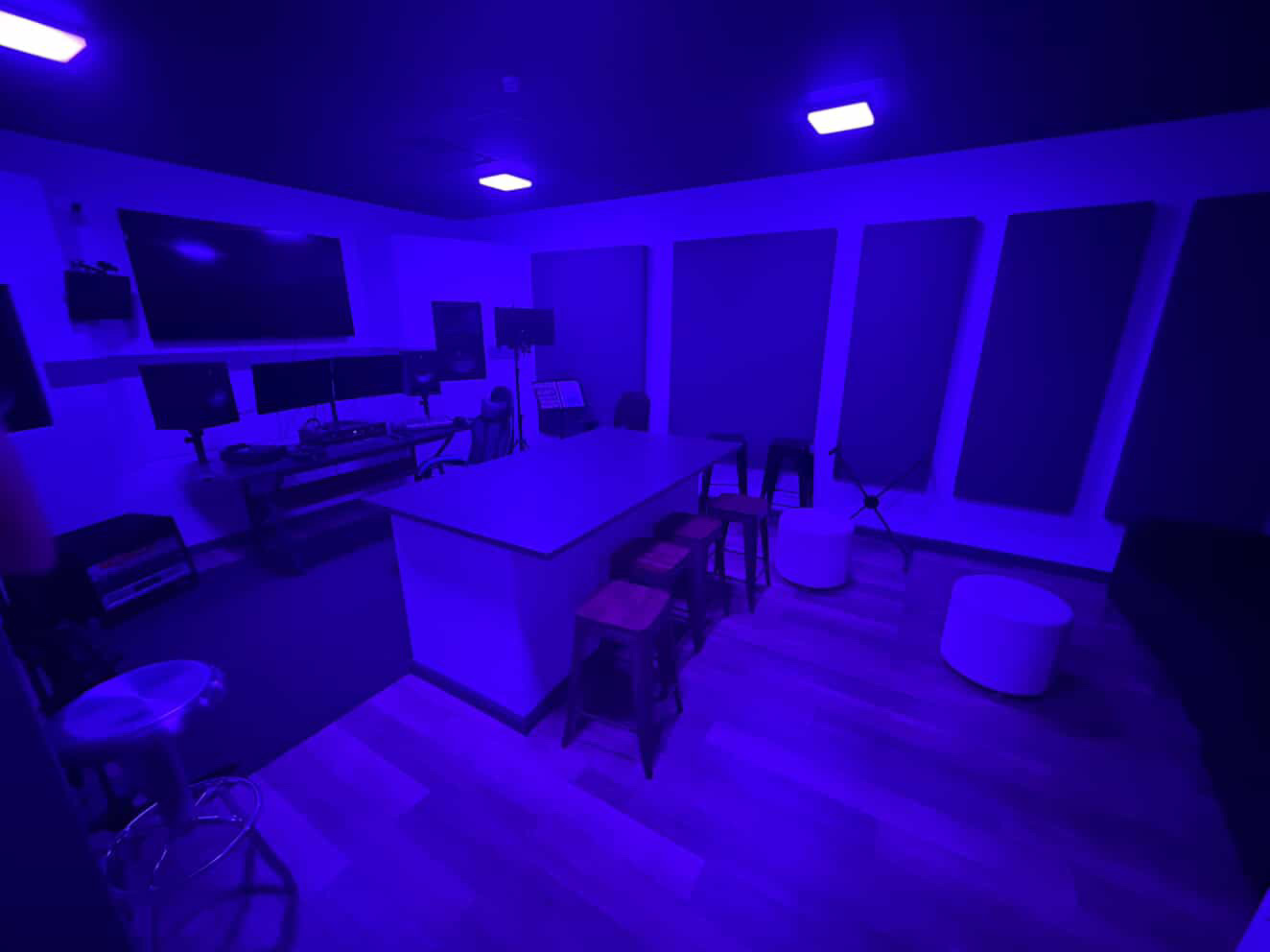 Room with a large table, chairs, and a TV, illuminated by blue lighting, with windows on one wall.