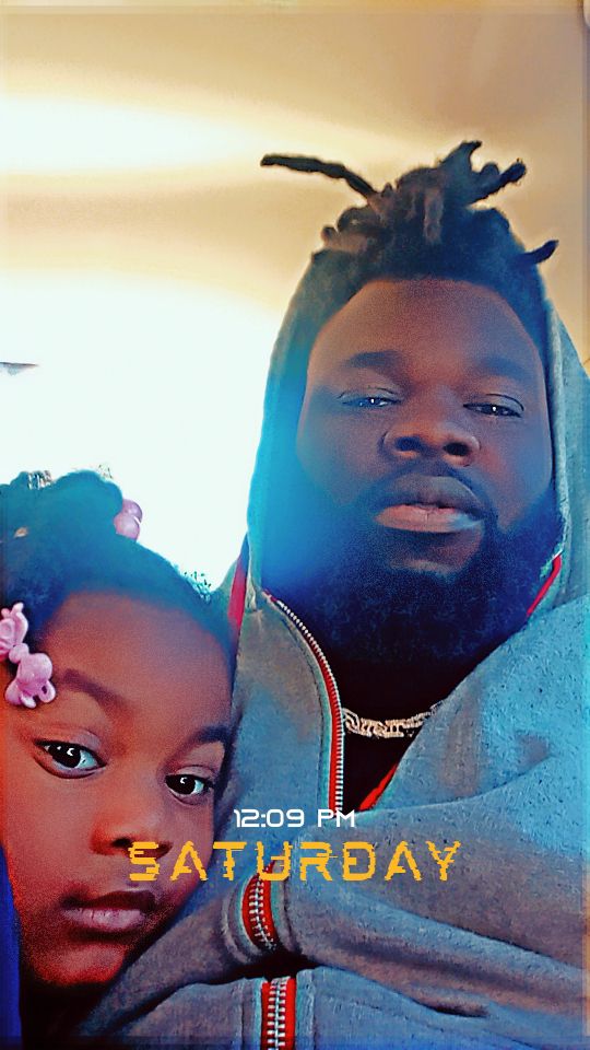 A man with dreadlocks and a young girl with flowers in her hair pose together indoors.