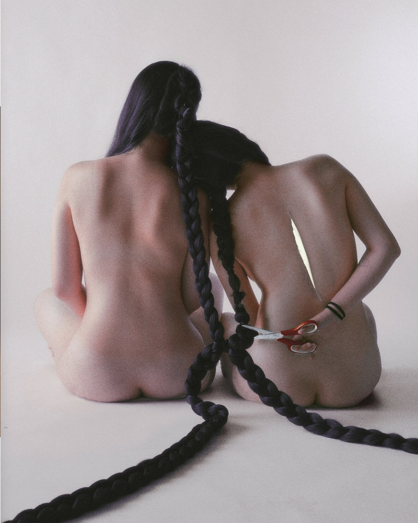 Two topless women with long braided hair sit on the floor, facing away from the camera, against a plain background.