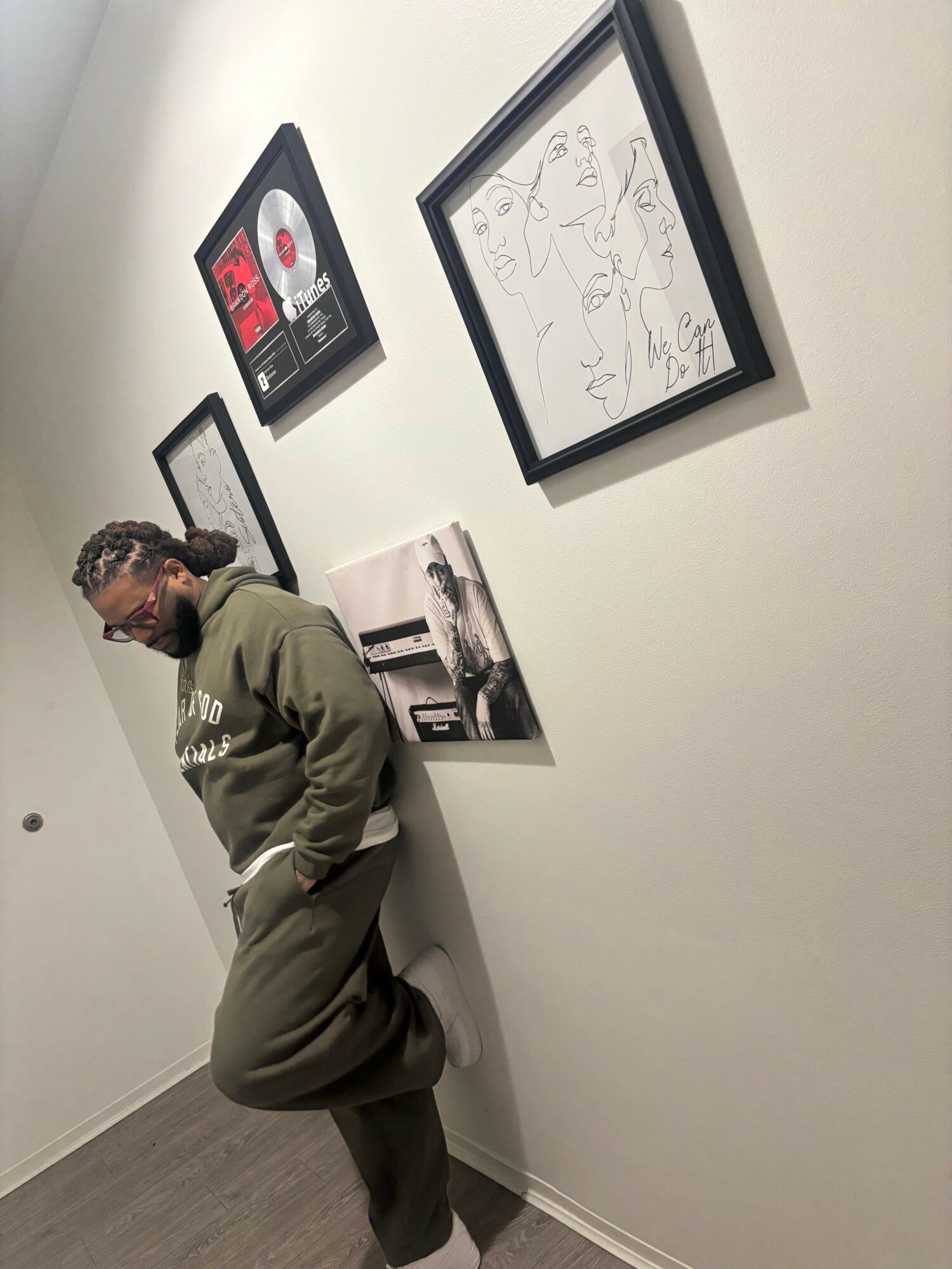 Man with gray hair and glasses leaning against a white wall with framed pictures, wearing a green jacket and dark pants.