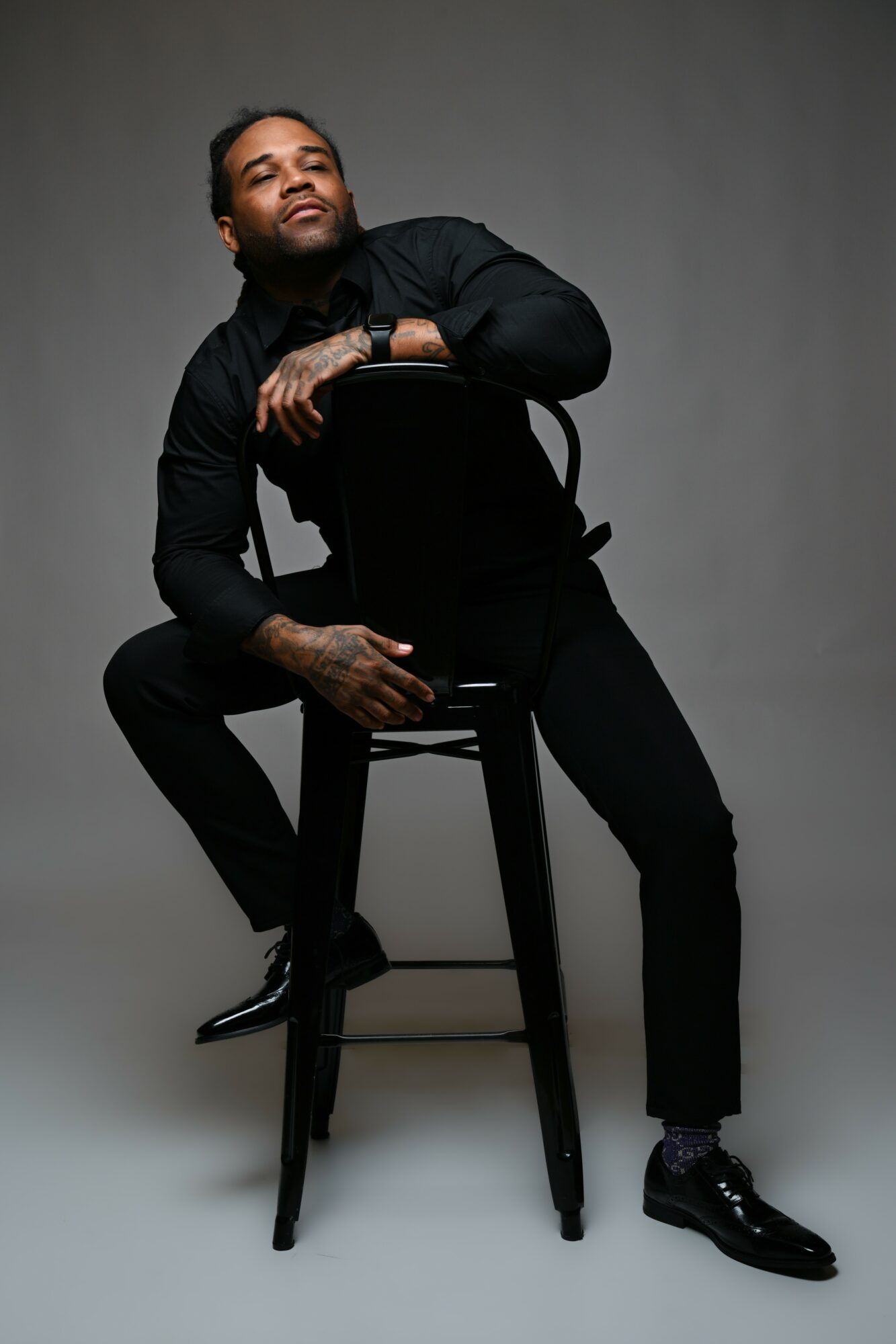 Person sitting on a chair, leaning on the backrest, wearing a black outfit, smiling, against a plain gray background.